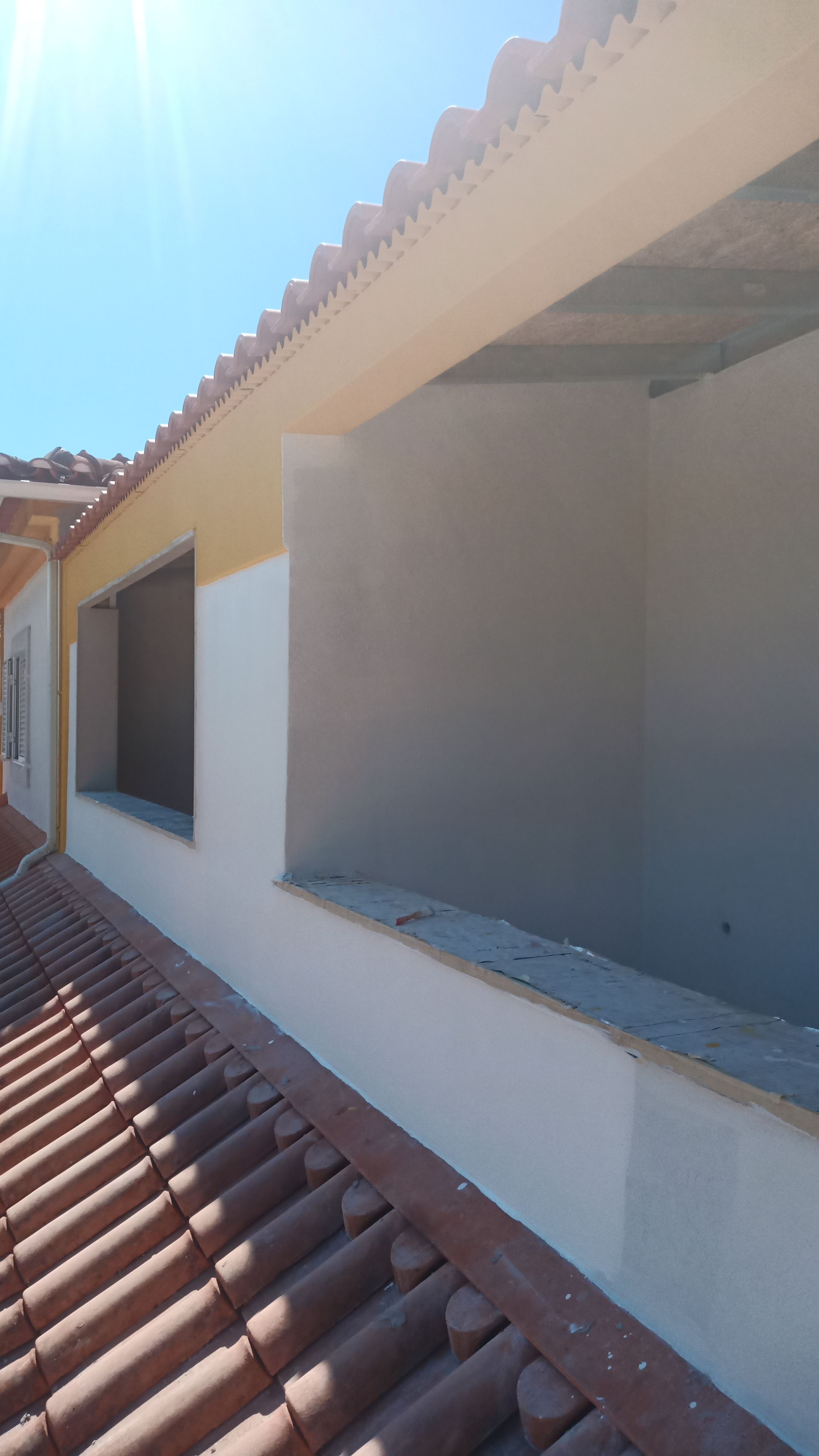 Exterior de um edifício com janela, paredes brancas, telhas laranja e céu ensolarado.