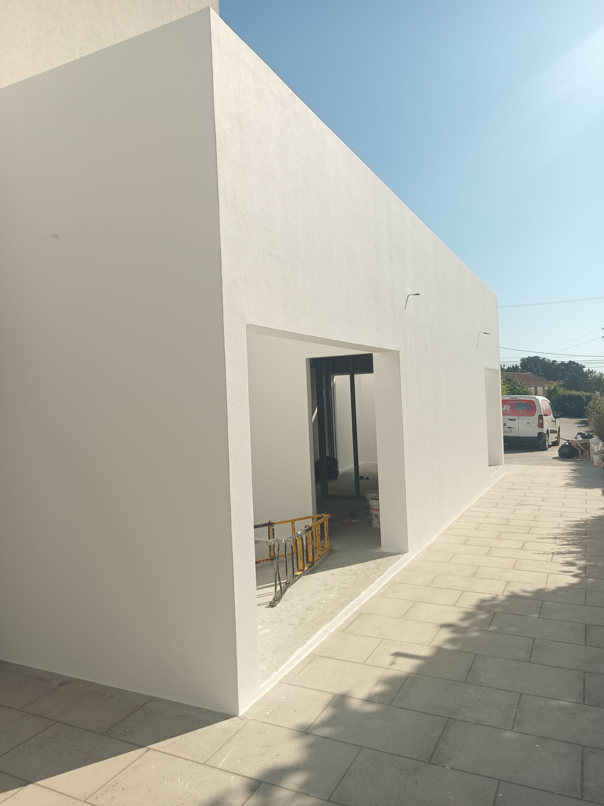 Prédio branco com porta aberta em calçada de pedra; rua com veículos estacionados visível ao fundo.