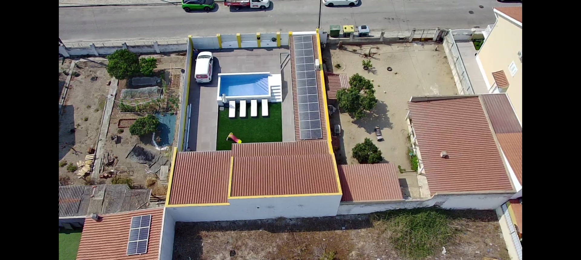 Vista aérea de uma propriedade com piscina, jardim e estacionamento junto a uma estrada.
