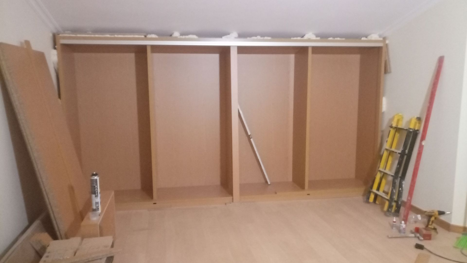 Um armário parcialmente construído com quatro compartimentos, em tom castanho claro, encostado a uma parede branca e piso de madeira clara.