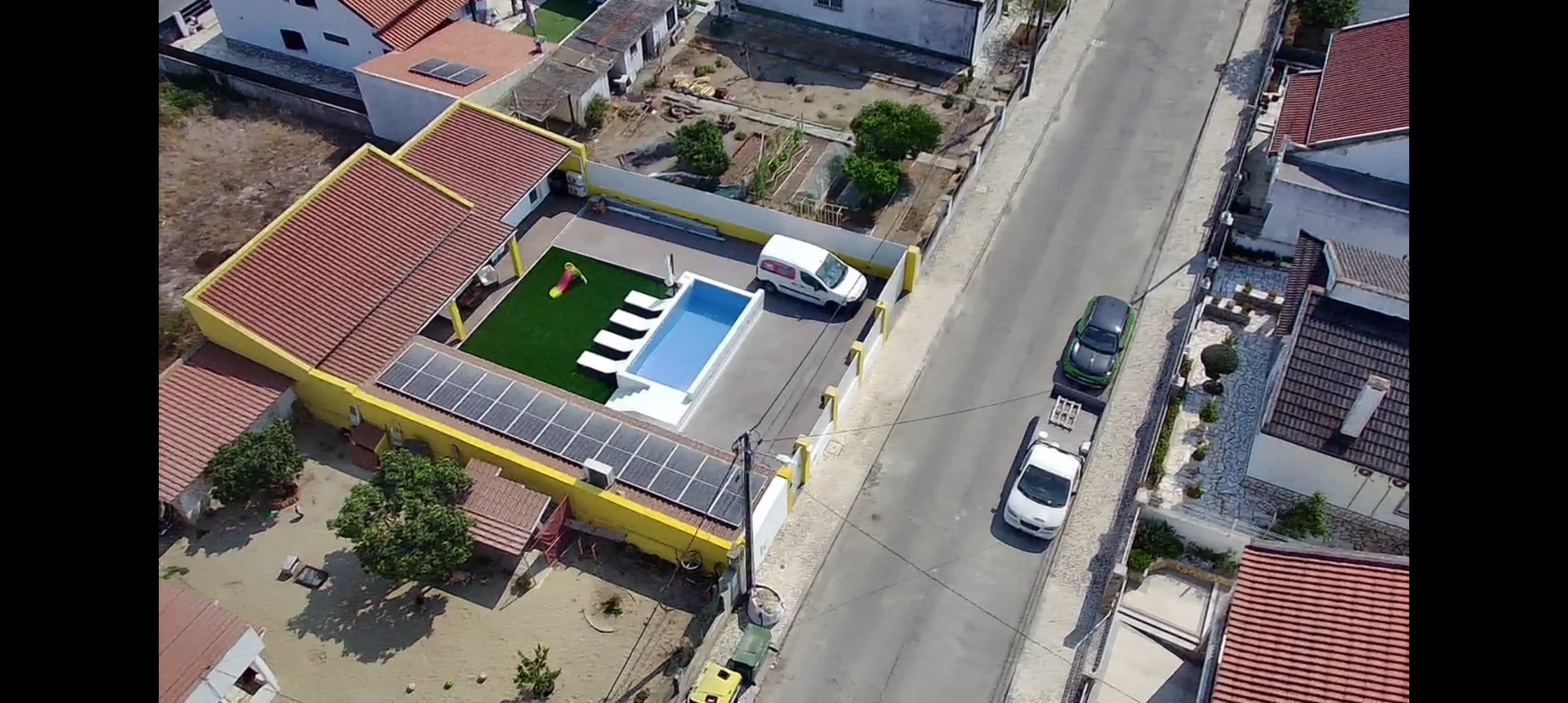 Vista aérea de um prédio amarelo com piscina, painéis solares e uma rua com carros.