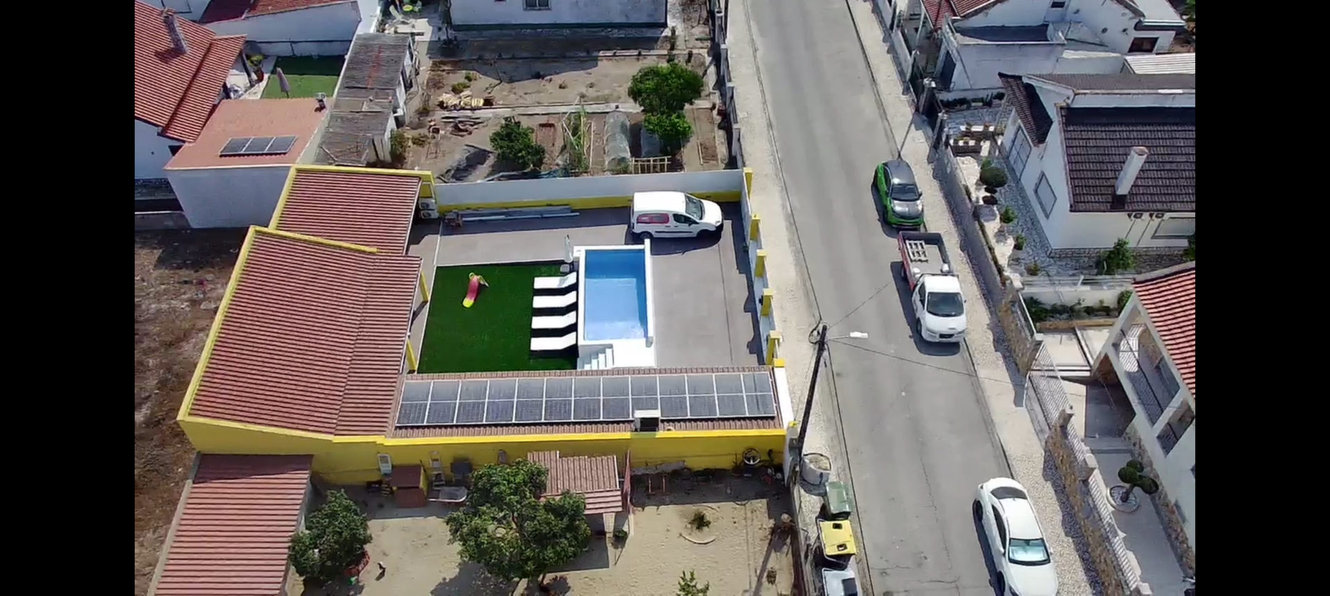 Vista aérea de um prédio amarelo com piscina, uma van branca e painéis solares ao longo de uma rua com carros.