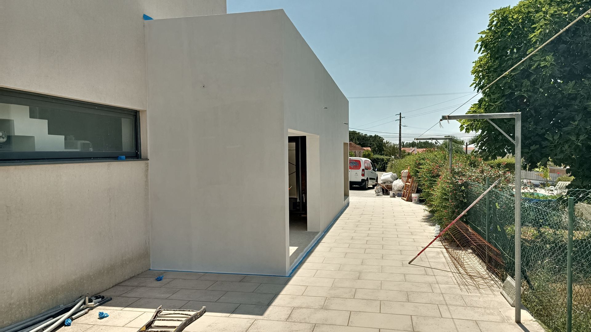 Vista exterior de um edifício branco com uma extensão inacabada, caminho ao lado, dia ensolarado.