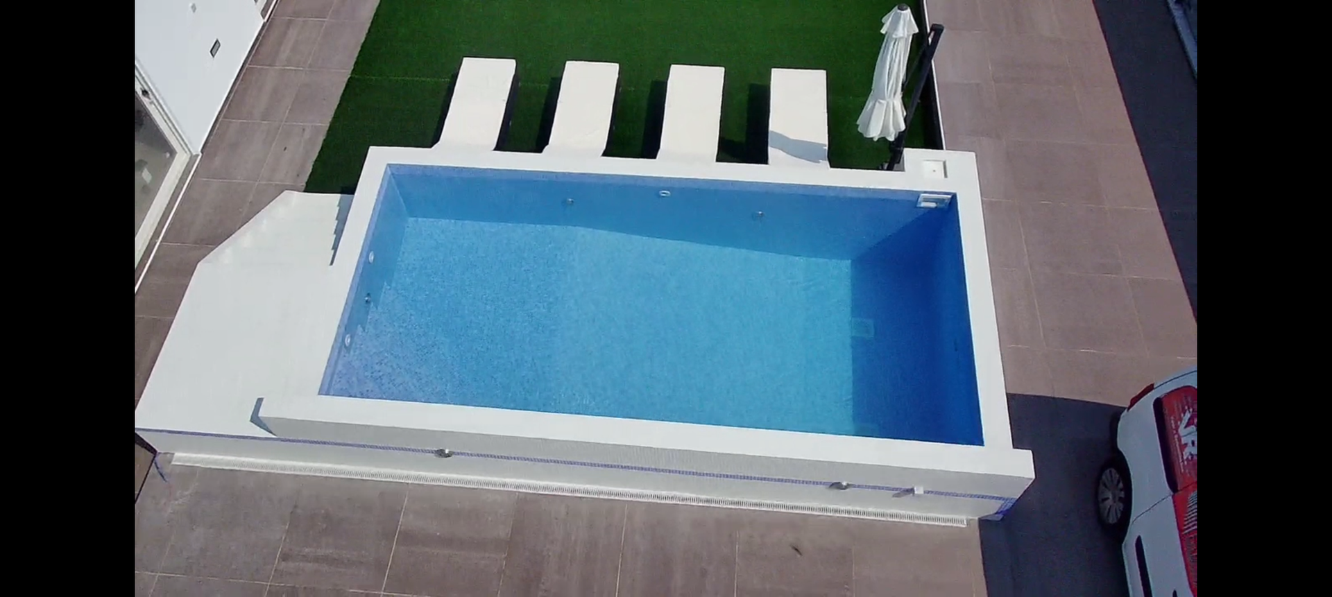 Vista aérea de uma piscina retangular azul rodeada por concreto branco. Espreguiçadeiras e grama artificial estão próximas.