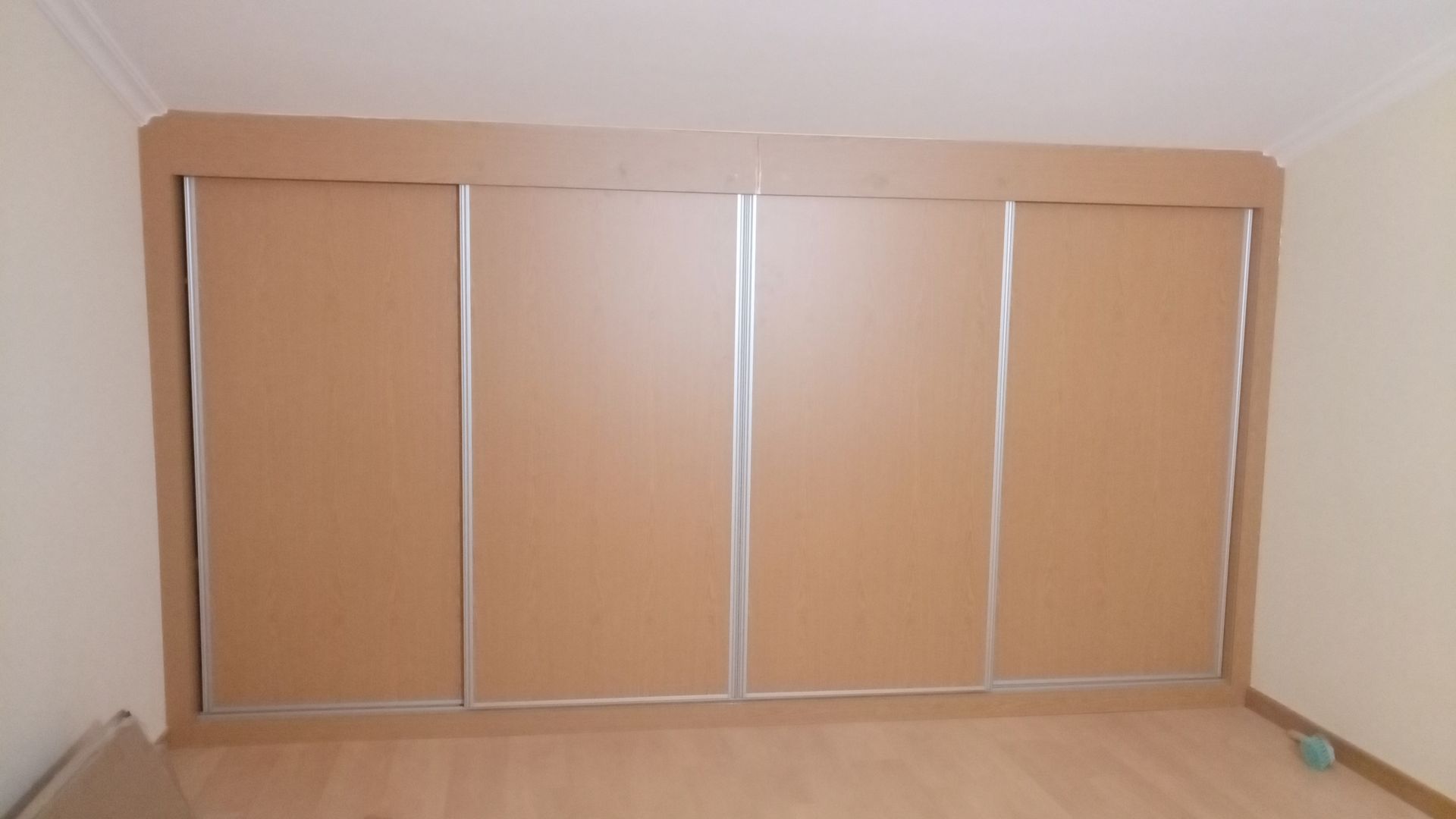 Armário embutido com quatro portas de correr, painéis marrons e detalhes prateados, em uma parede de um quarto com piso de madeira clara.