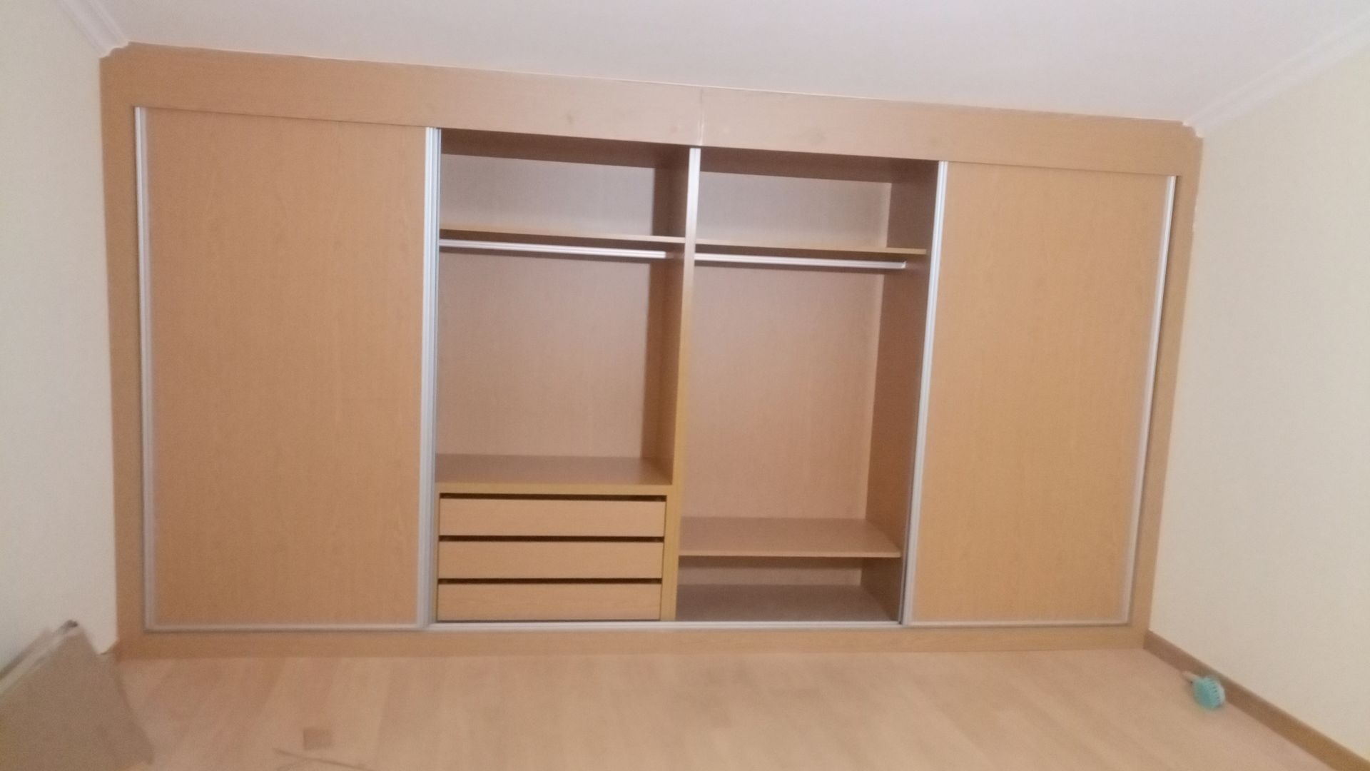 Um armário embutido marrom claro com portas de correr, prateleiras abertas, varões para pendurar roupas e gavetas, em um quarto com piso de madeira.