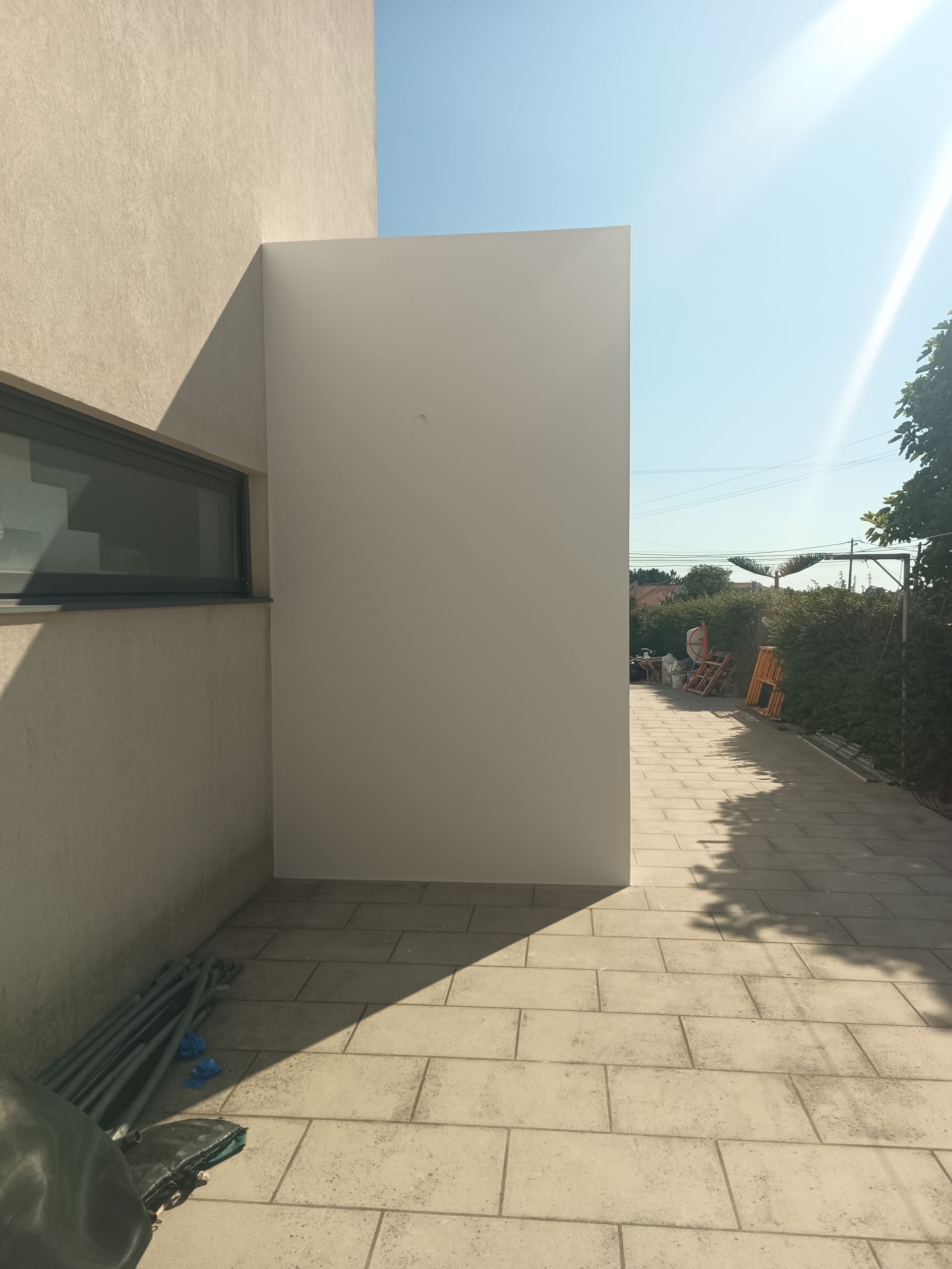 Parede branca ao lado de uma casa com janela, caminho externo ensolarado.