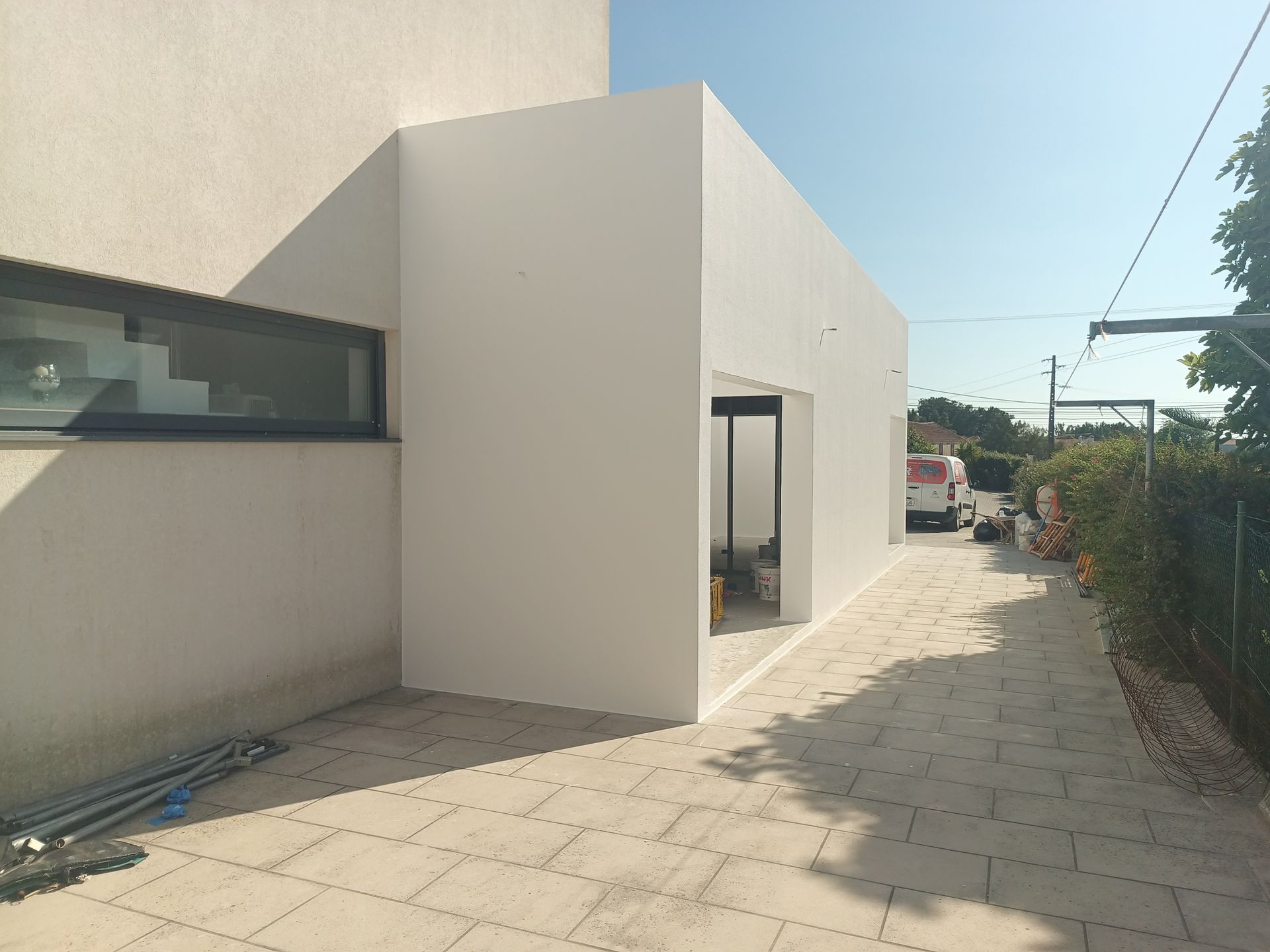 Fachada branca de um prédio com entrada lateral, caminho pavimentado e veículo estacionado. Dia ensolarado.
