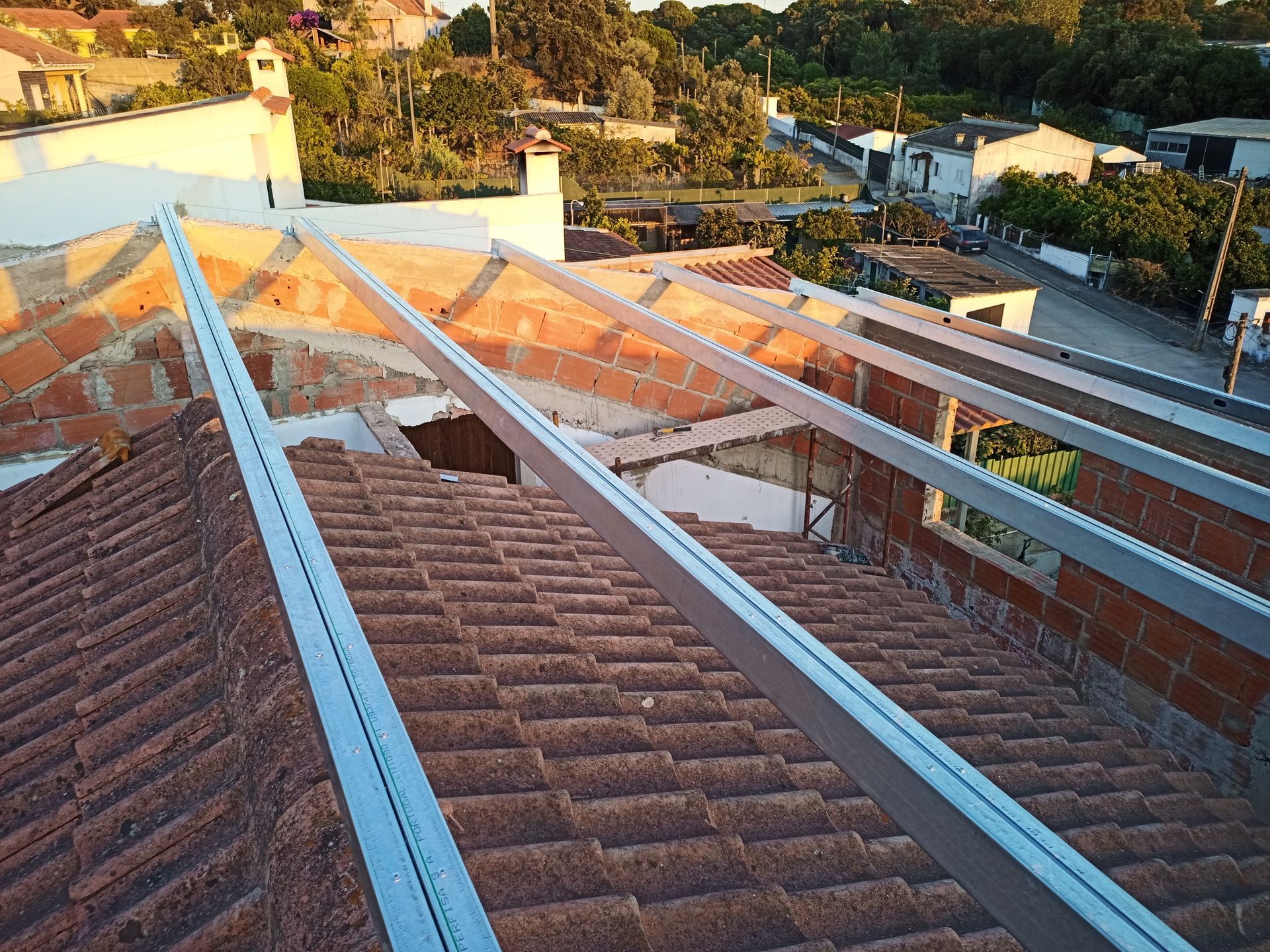 Vigas metálicas dispostas sobre um telhado de telhas vermelhas, provavelmente para a instalação de painéis solares, com uma paisagem urbana ao fundo.