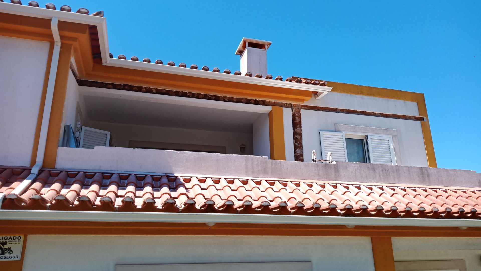 Prédio com detalhes em laranja, telhado de telha vermelha, varanda, chaminé e céu azul.