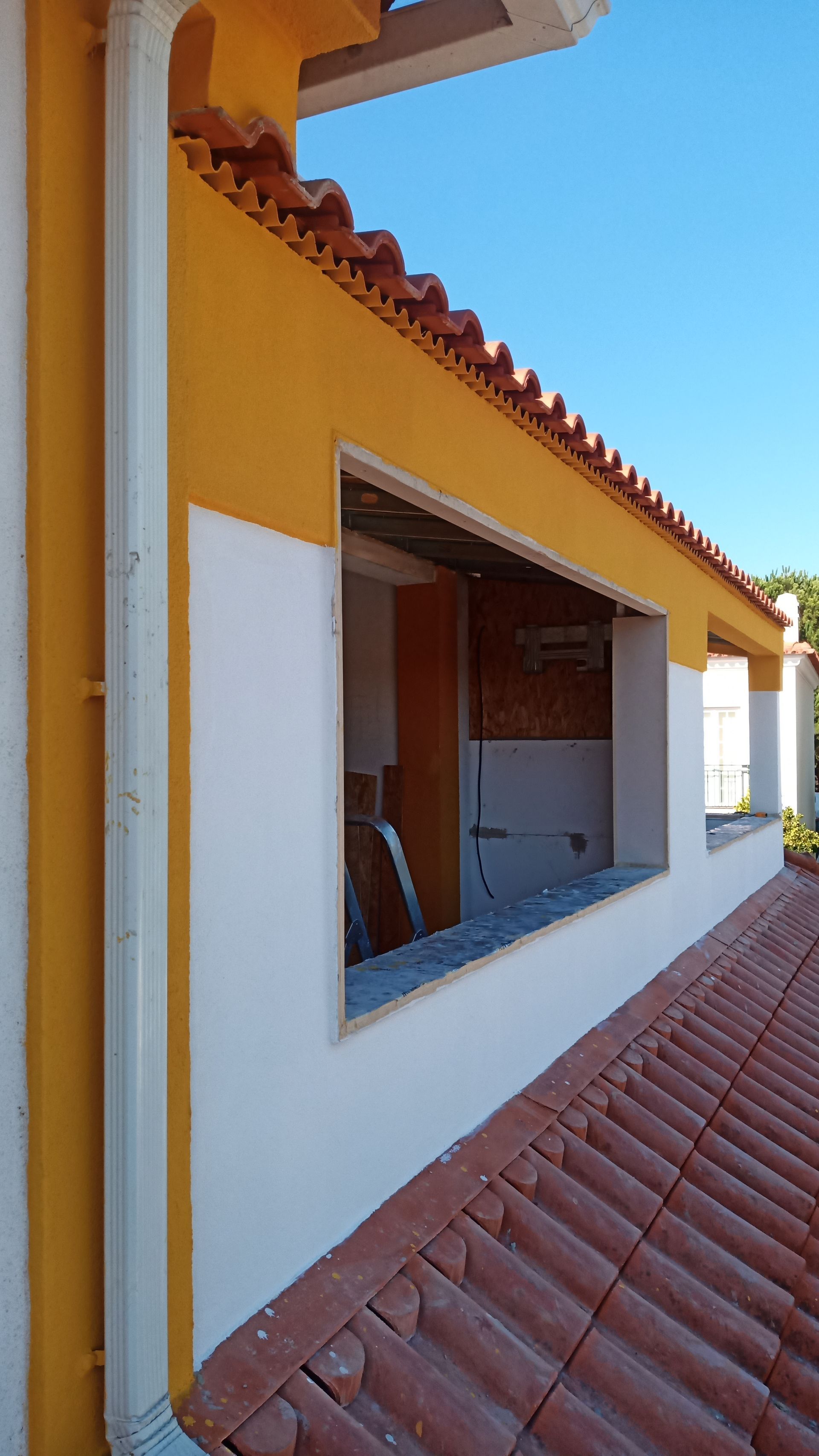 Parede externa com janela aberta, pintura amarela e branca, telhado de telha vermelha, céu limpo.