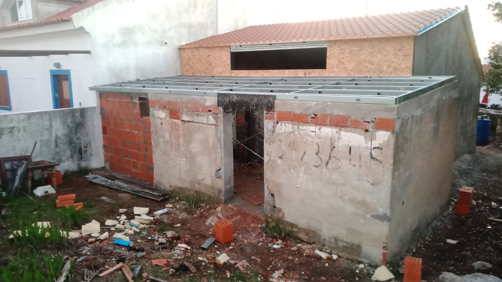 Prédio em construção com tijolos e compensado aparentes, vista externa, terreno de terra batida, iluminação natural.