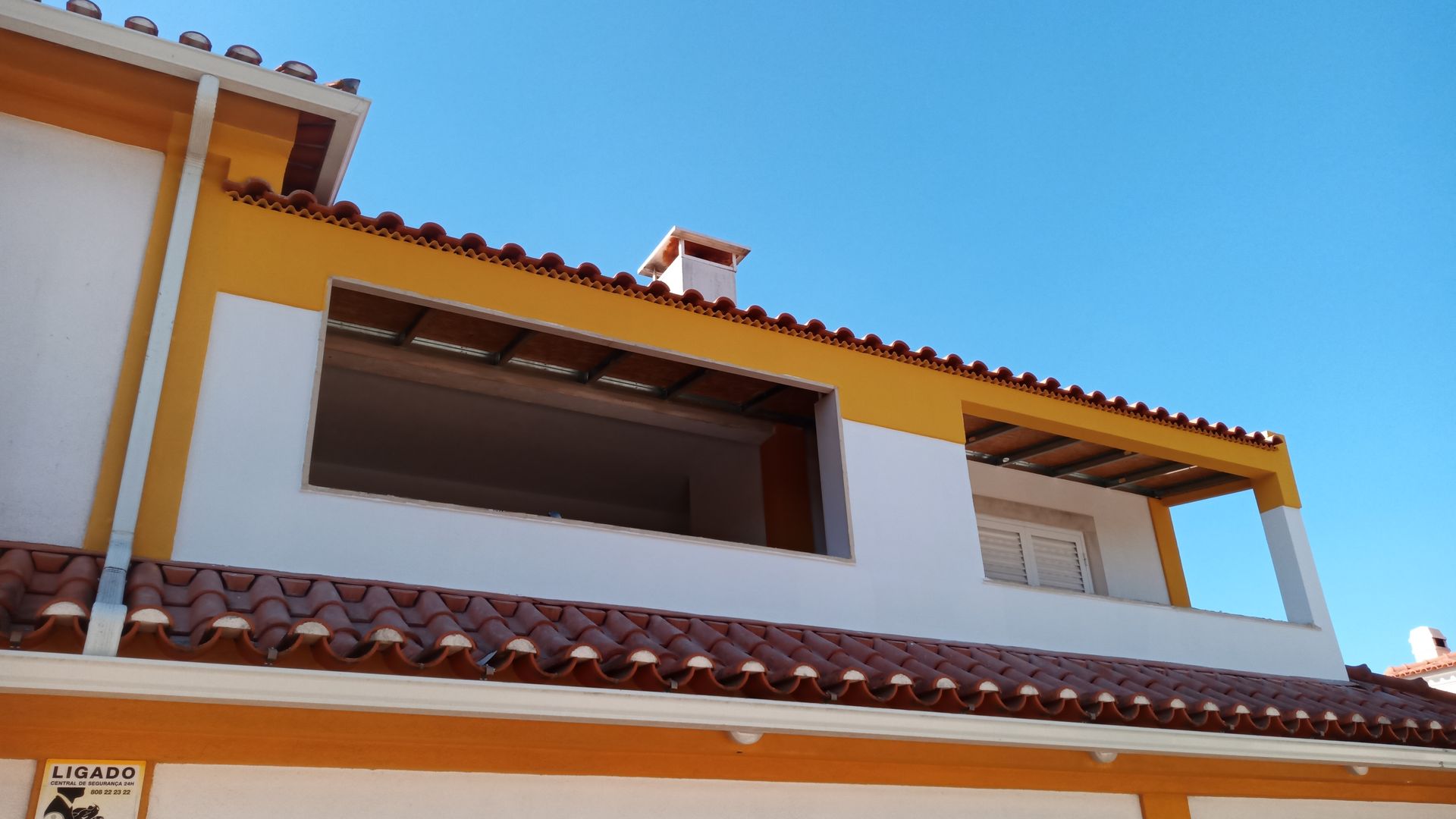 Prédio branco e amarelo com telhado de telhas vermelhas contra um céu azul claro. Janela aberta no segundo andar.