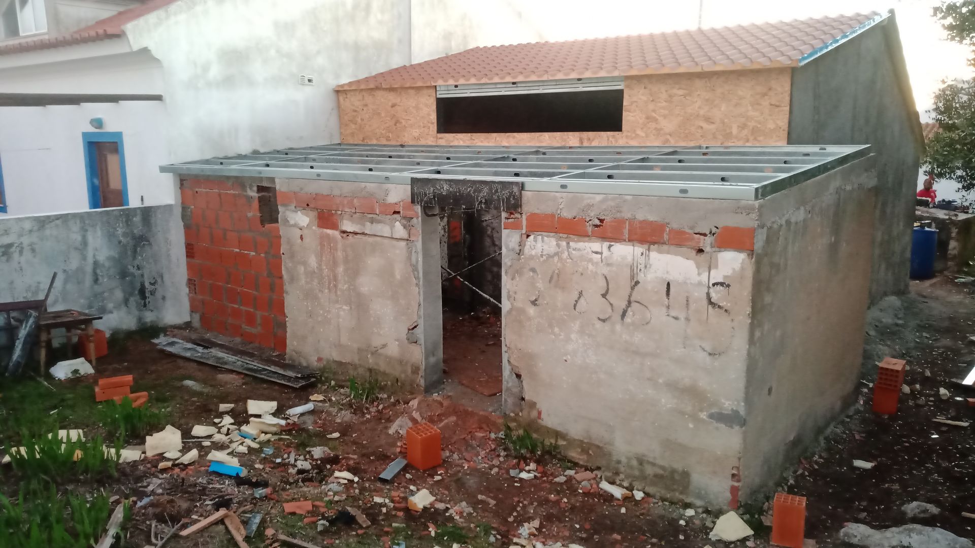 Construção de uma pequena estrutura de tijolo e concreto com porta aberta e telhado inacabado. Ambiente externo.