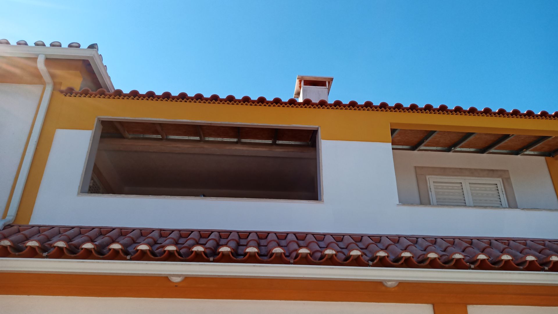 Prédio com telhado de telhas laranja, paredes brancas e amarelas, uma janela e céu azul.