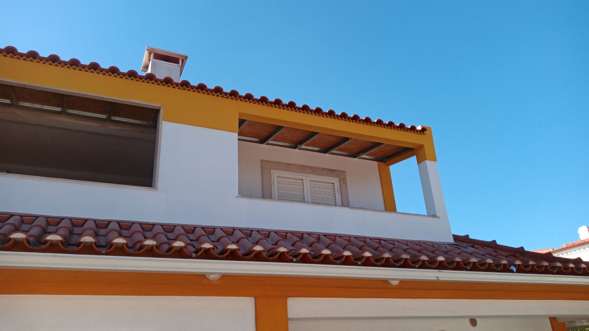 Casa branca com detalhes em laranja, telhado de telha vermelha e varanda contra um céu azul.