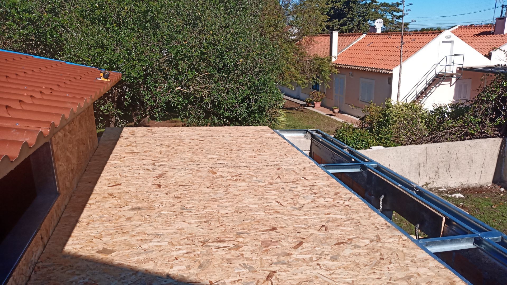 Cobertura de OSB em construção, ao lado de um telhado de telhas vermelhas e outros edifícios, em um dia ensolarado.