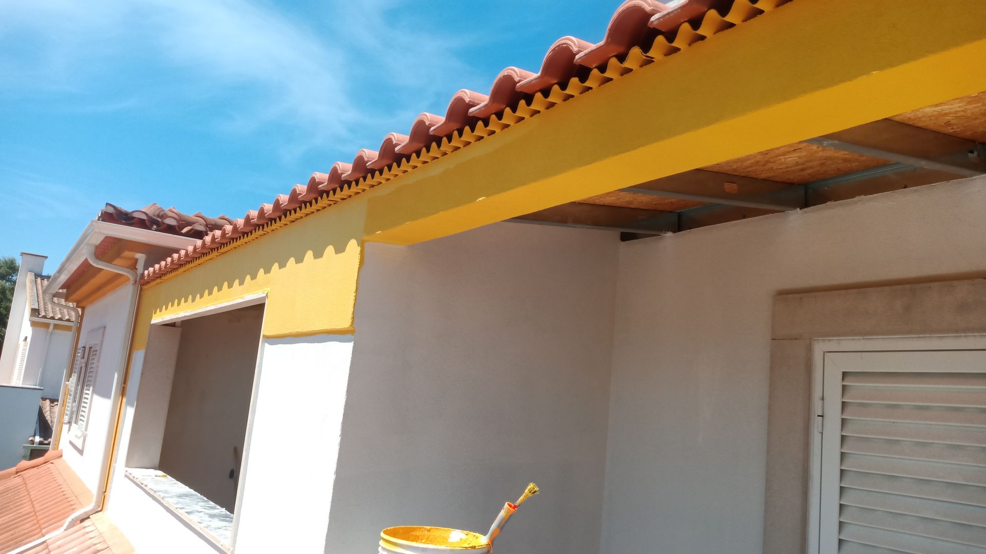 Vista exterior de um edifício com telhado de telhas vermelhas, detalhes em amarelo e paredes brancas, contra um céu azul.