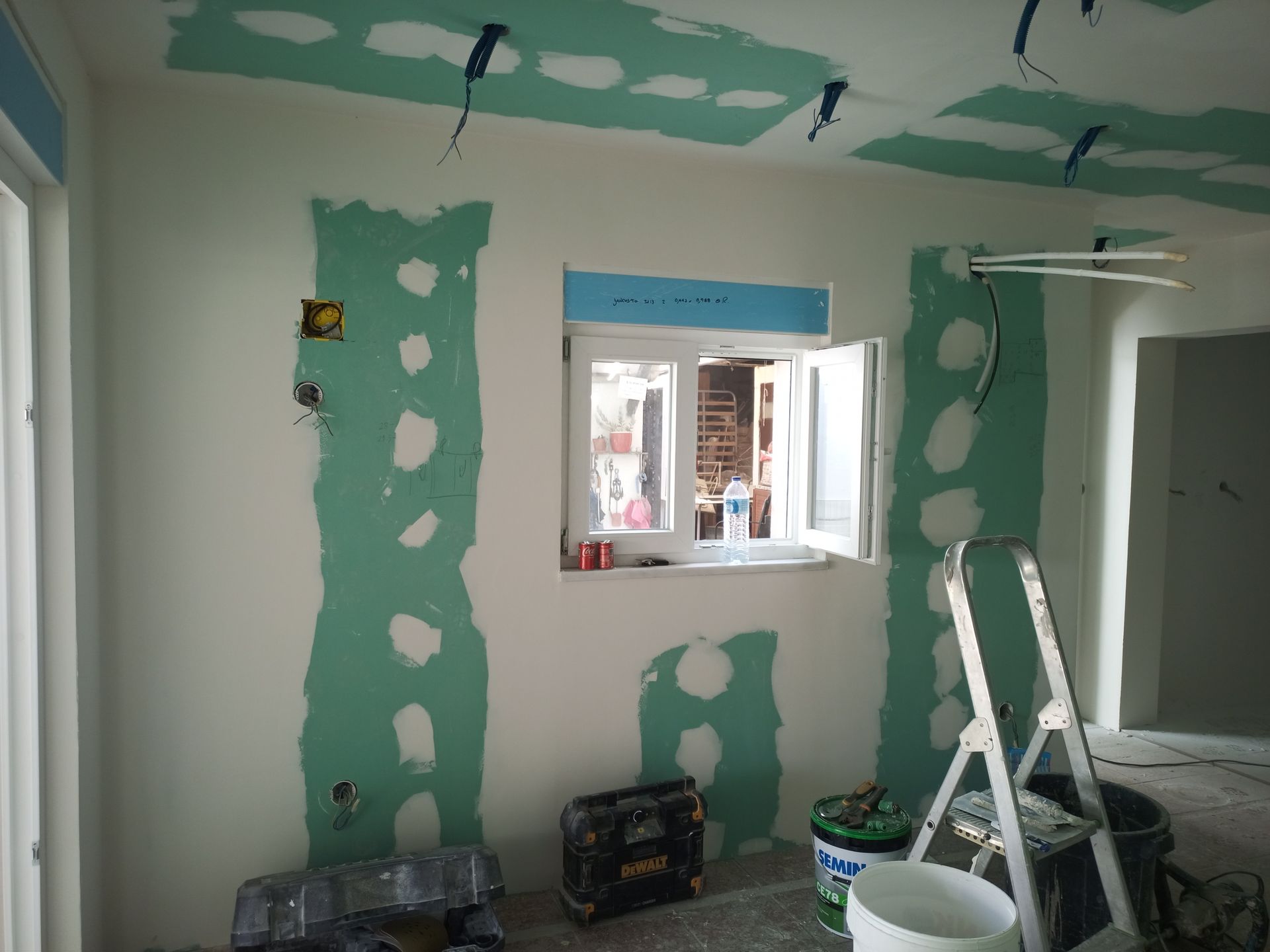 Quarto em construção: drywall verde, fiação elétrica, paredes brancas, janela aberta e escada.