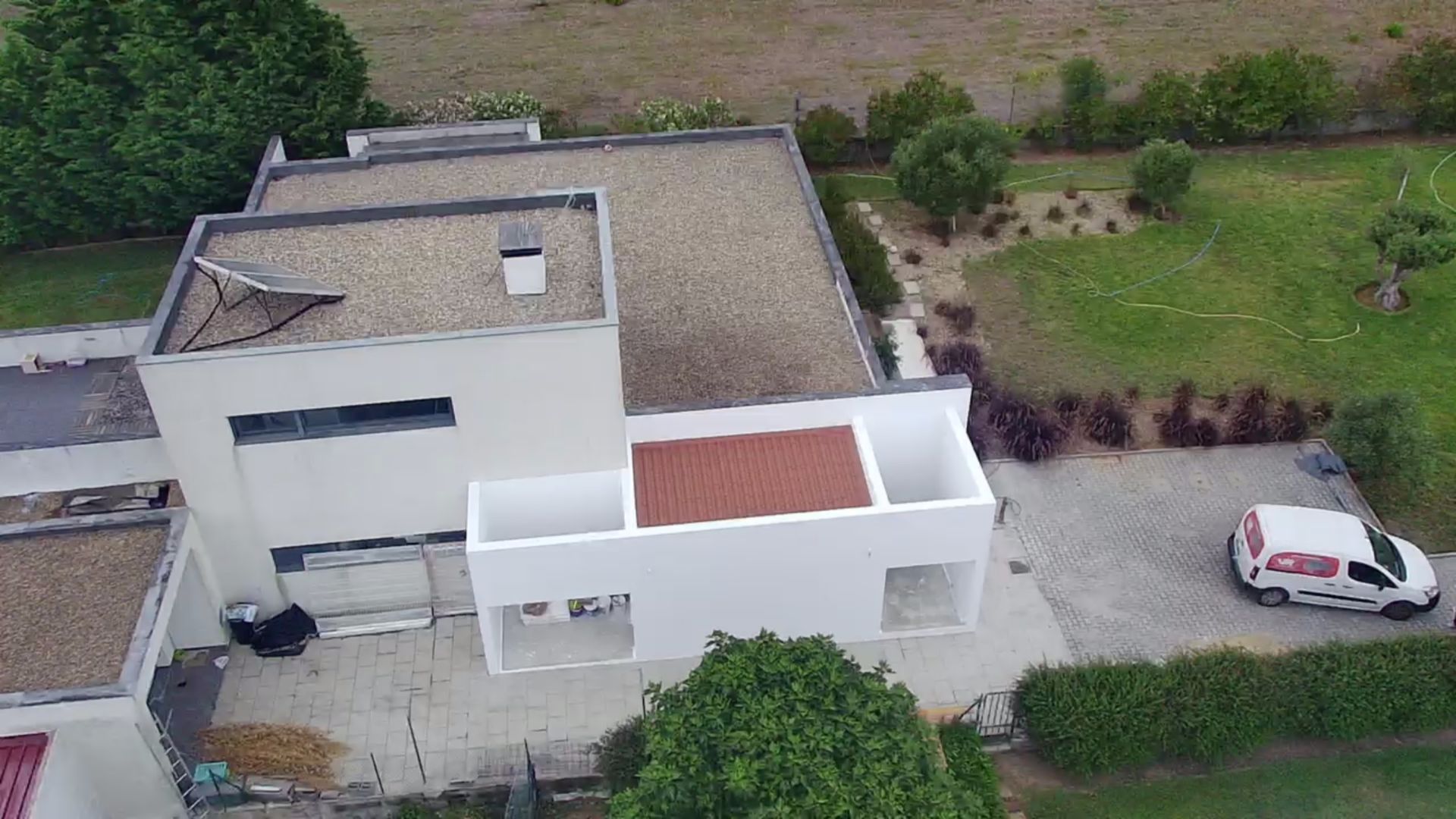 Casa moderna branca com telhados planos e detalhes em telha vermelha, van branca estacionada à direita e quintal verde.