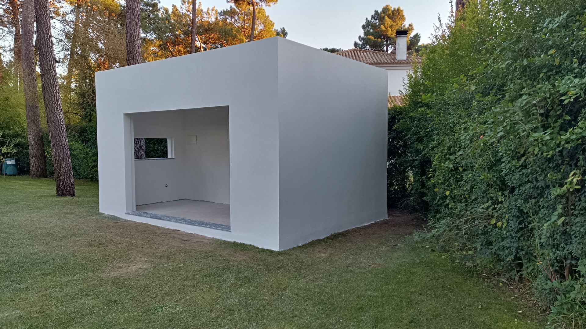 Estrutura branca em forma de cubo com uma porta aberta e uma pequena janela, situada num pátio gramado, rodeada por árvores e arbustos.