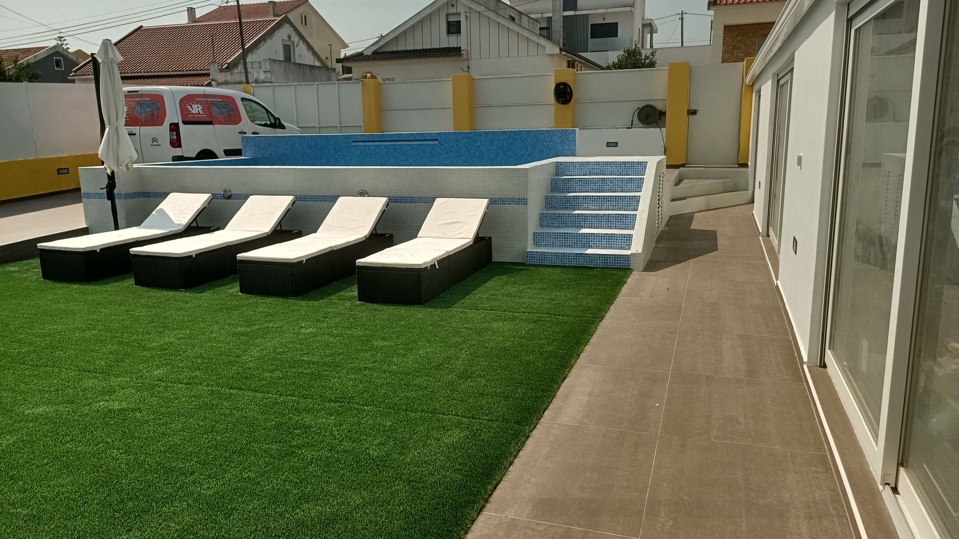 Uma piscina com espreguiçadeiras sobre grama artificial, ao lado de um prédio. Uma van branca e uma cerca ao fundo.