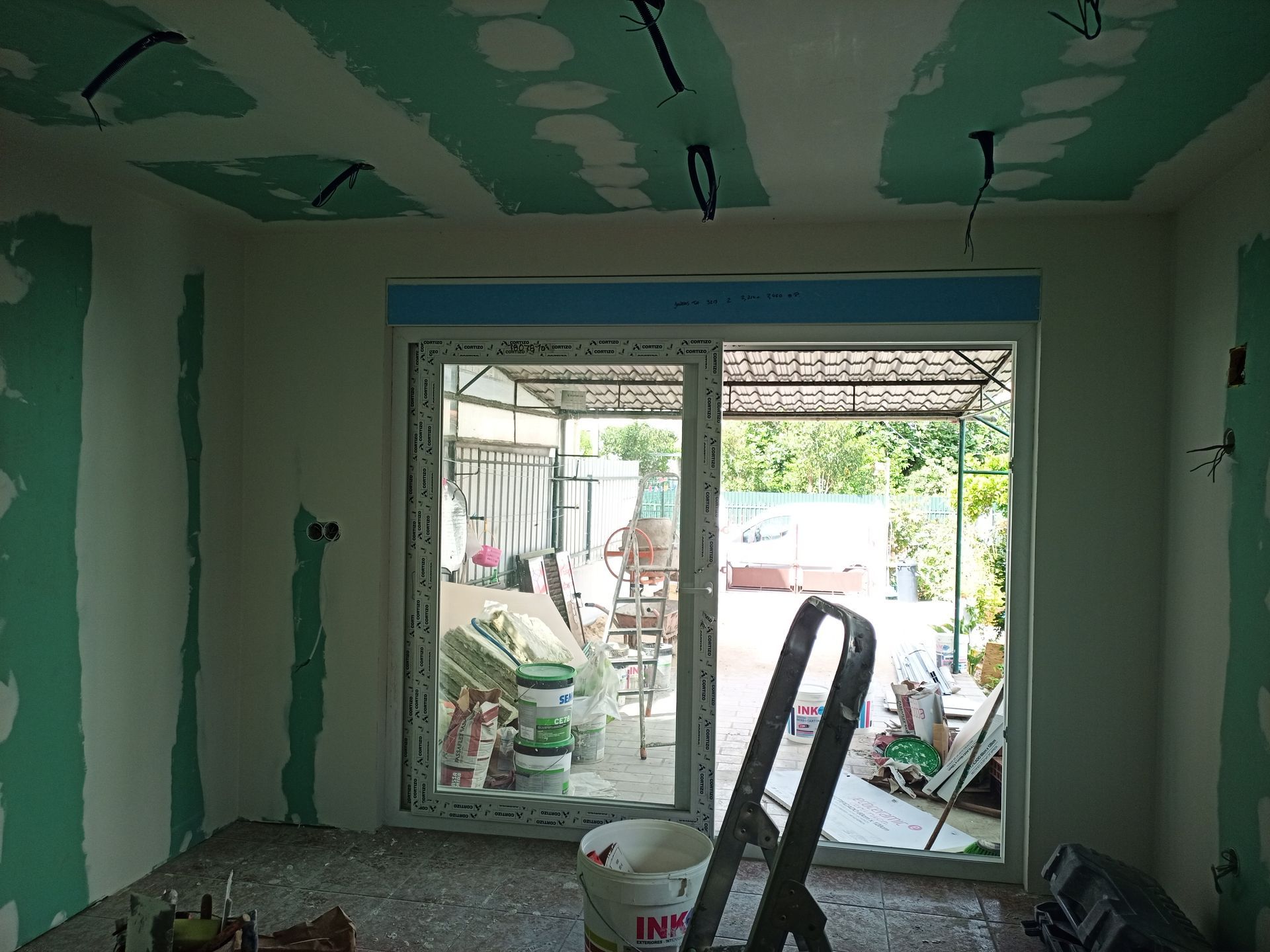 Quarto interno em construção, com drywall verde, porta de vidro deslizante e escada de mão.