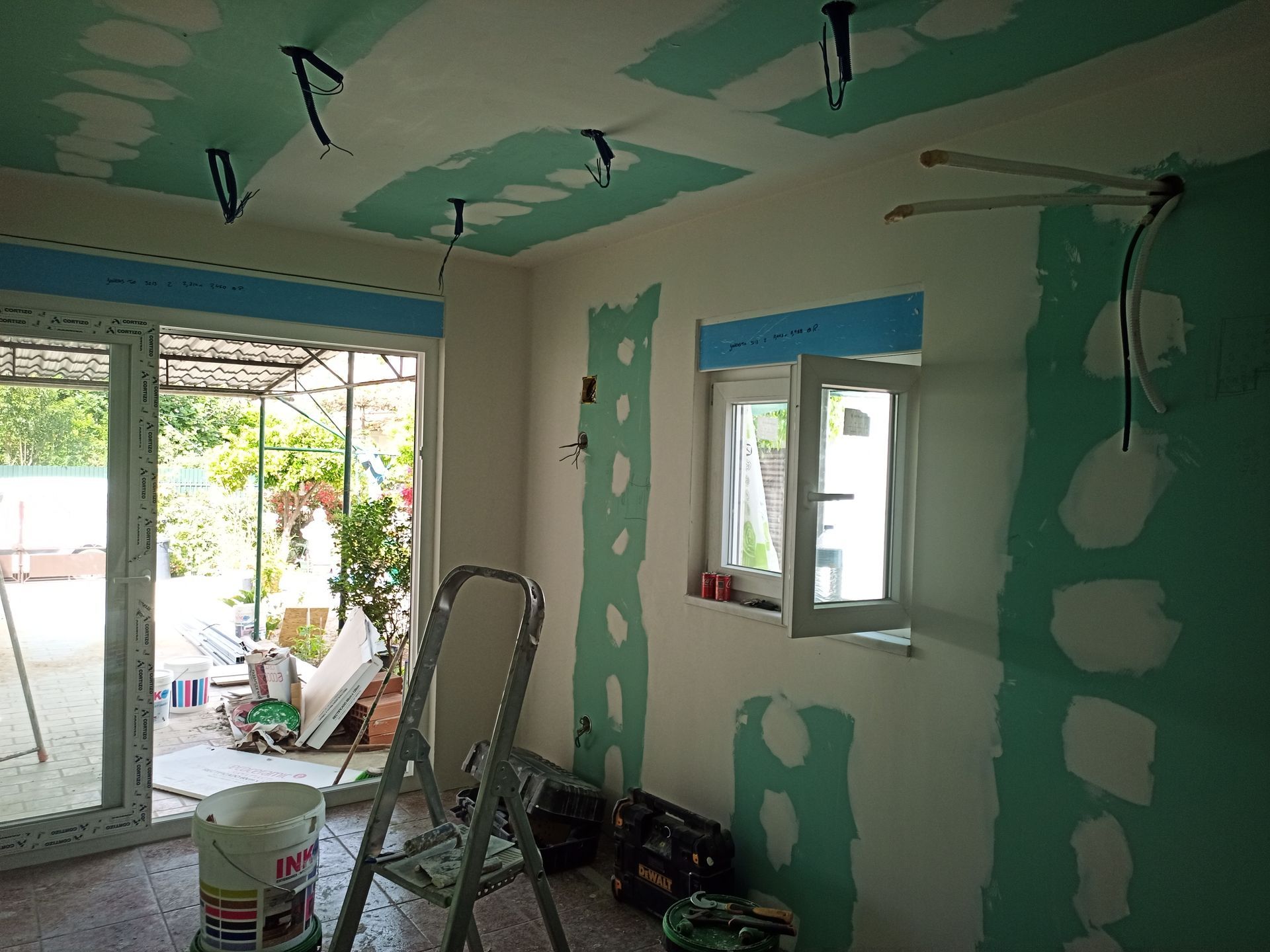 Quarto interior com drywall parcialmente acabado e fiação exposta. Uma escada e ferramentas estão presentes.