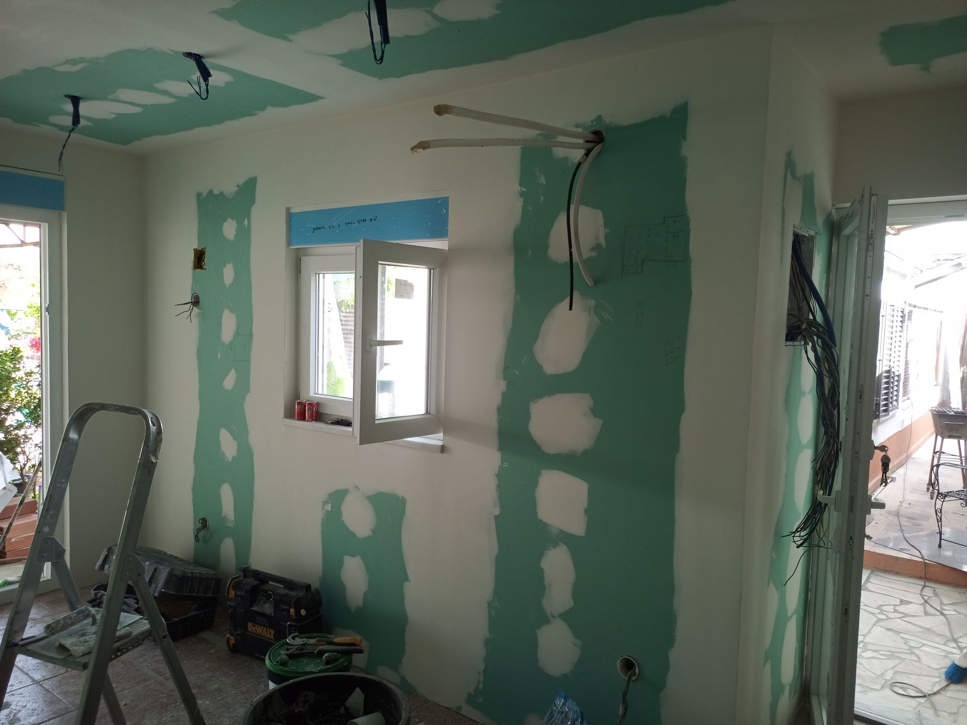 Interior em construção; instalação de drywall com fiação, janela e escada visíveis.