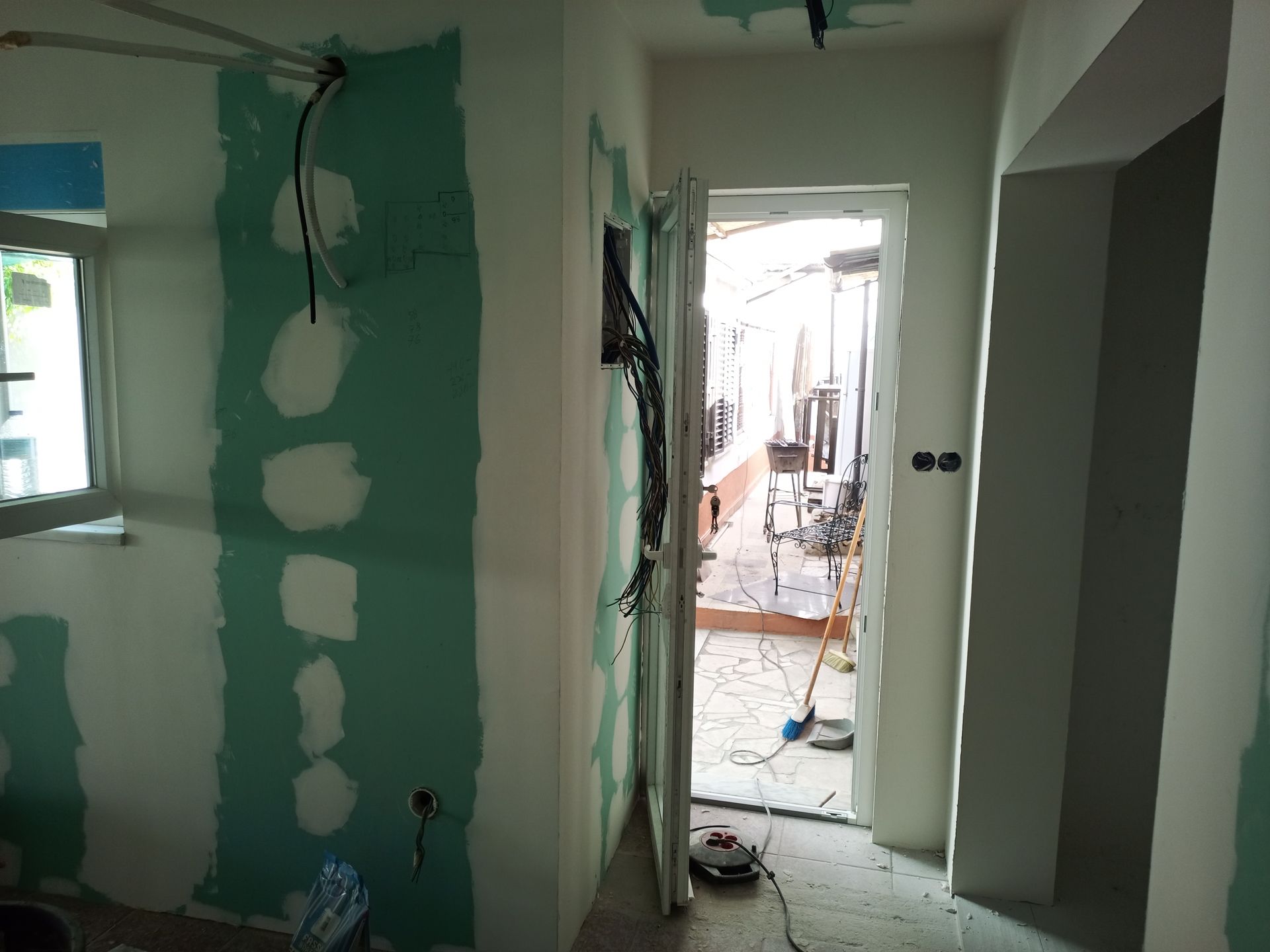 Vista interna de um cômodo em construção com fiação exposta, drywall verde e uma porta aberta.