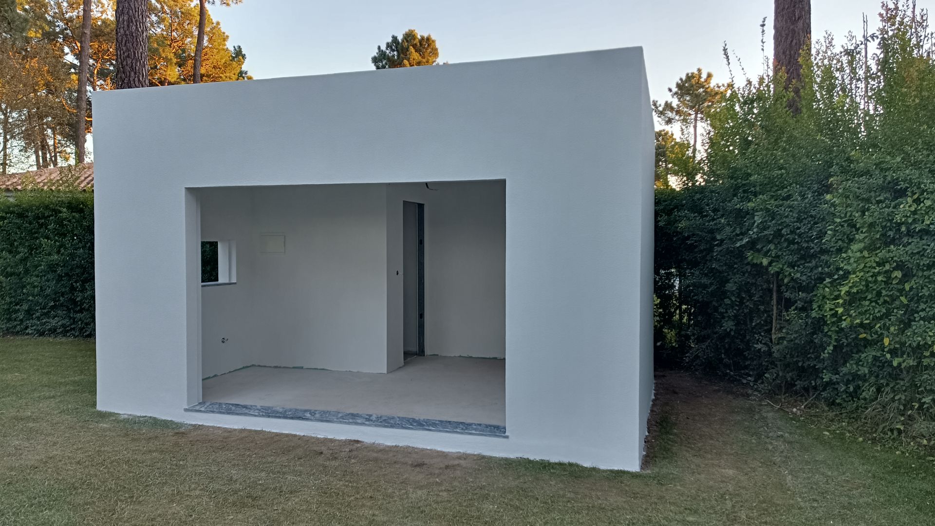 Estrutura cúbica branca com a fachada aberta e um cômodo interno, em uma área gramada.