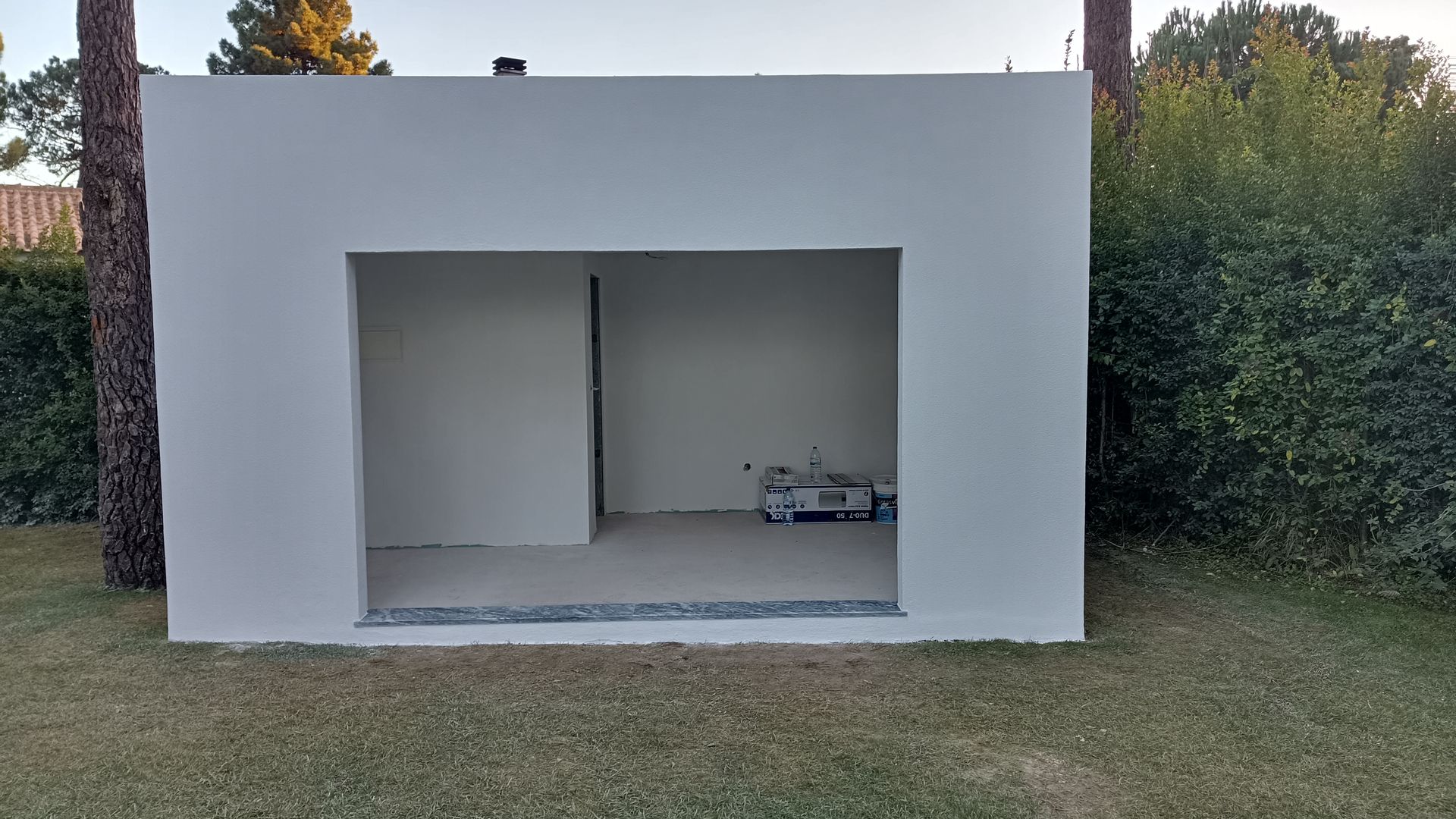 Estrutura quadrada branca com uma grande abertura para a grama. O interior revela um cômodo vazio.