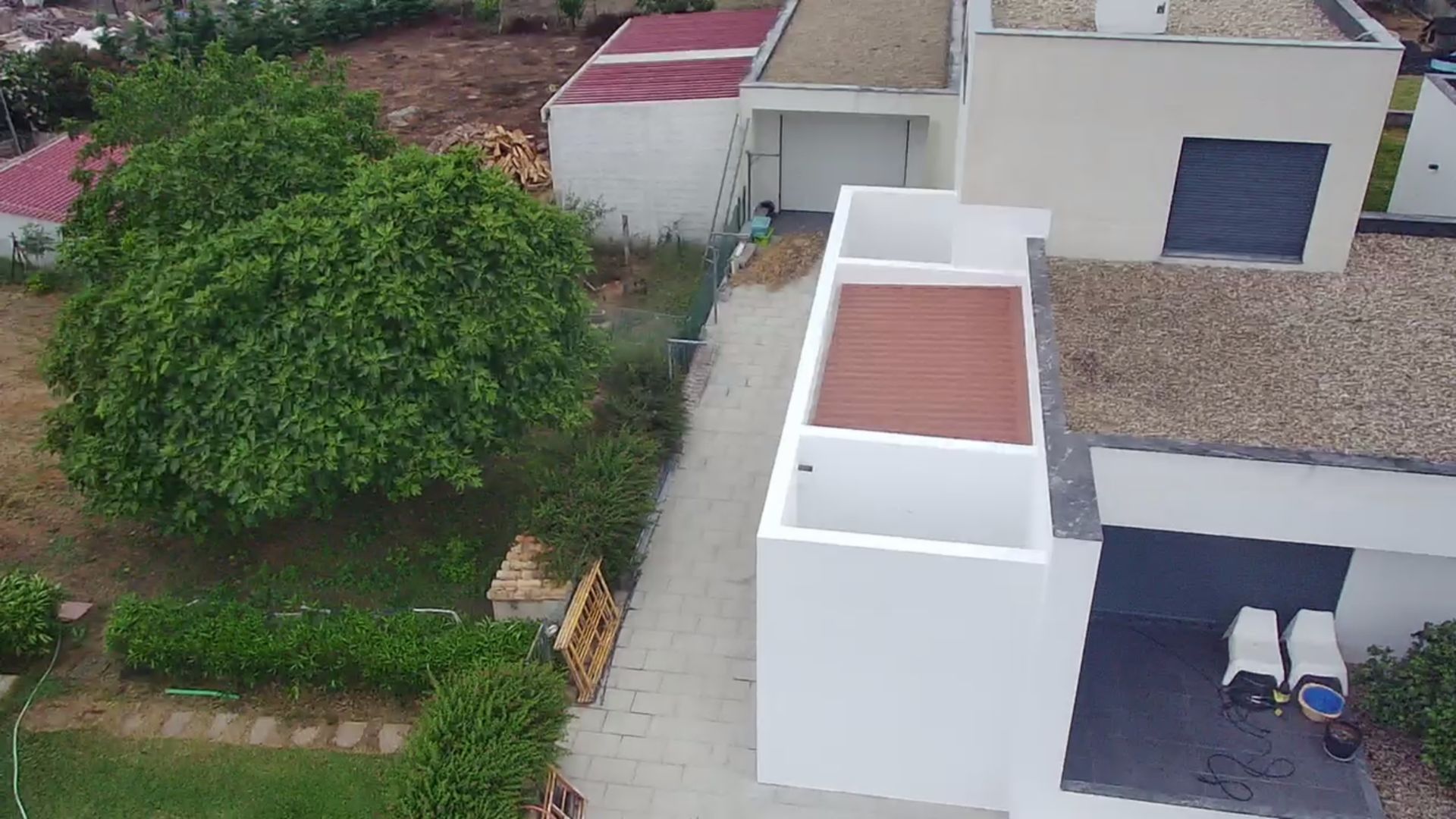 Vista aérea de uma casa moderna branca com uma passarela, rodeada por vegetação e outros edifícios.