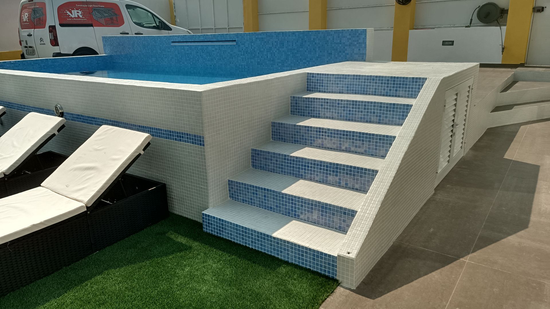 Piscina com azulejos azuis e degraus. Duas espreguiçadeiras estão dispostas na grama.