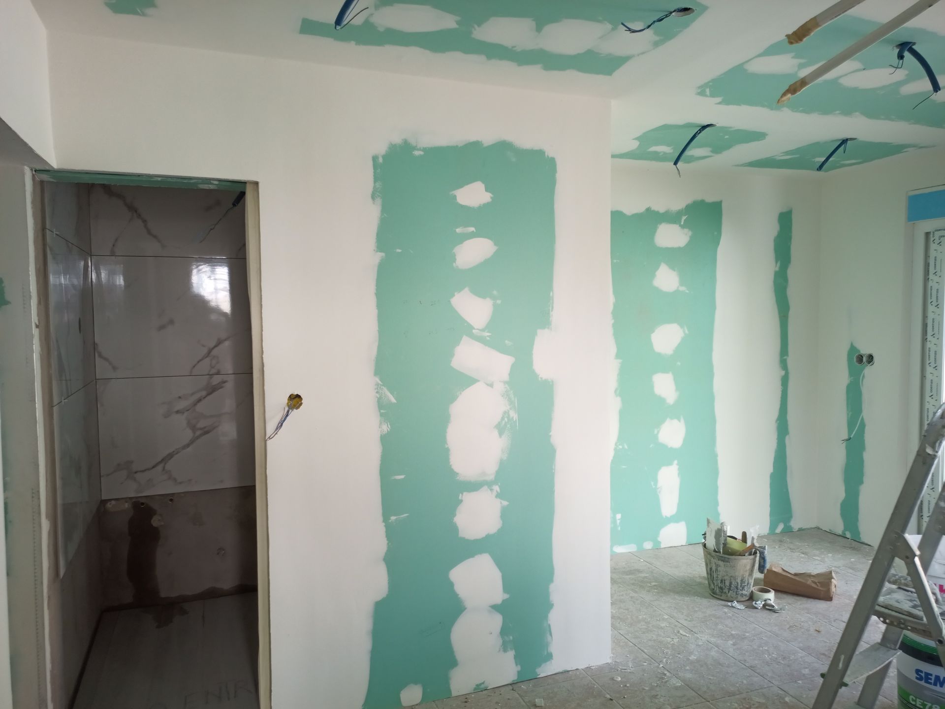 Vista interna de um cômodo em reforma. Paredes e teto revestidos com gesso acartonado verde; porta que dá acesso ao banheiro.
