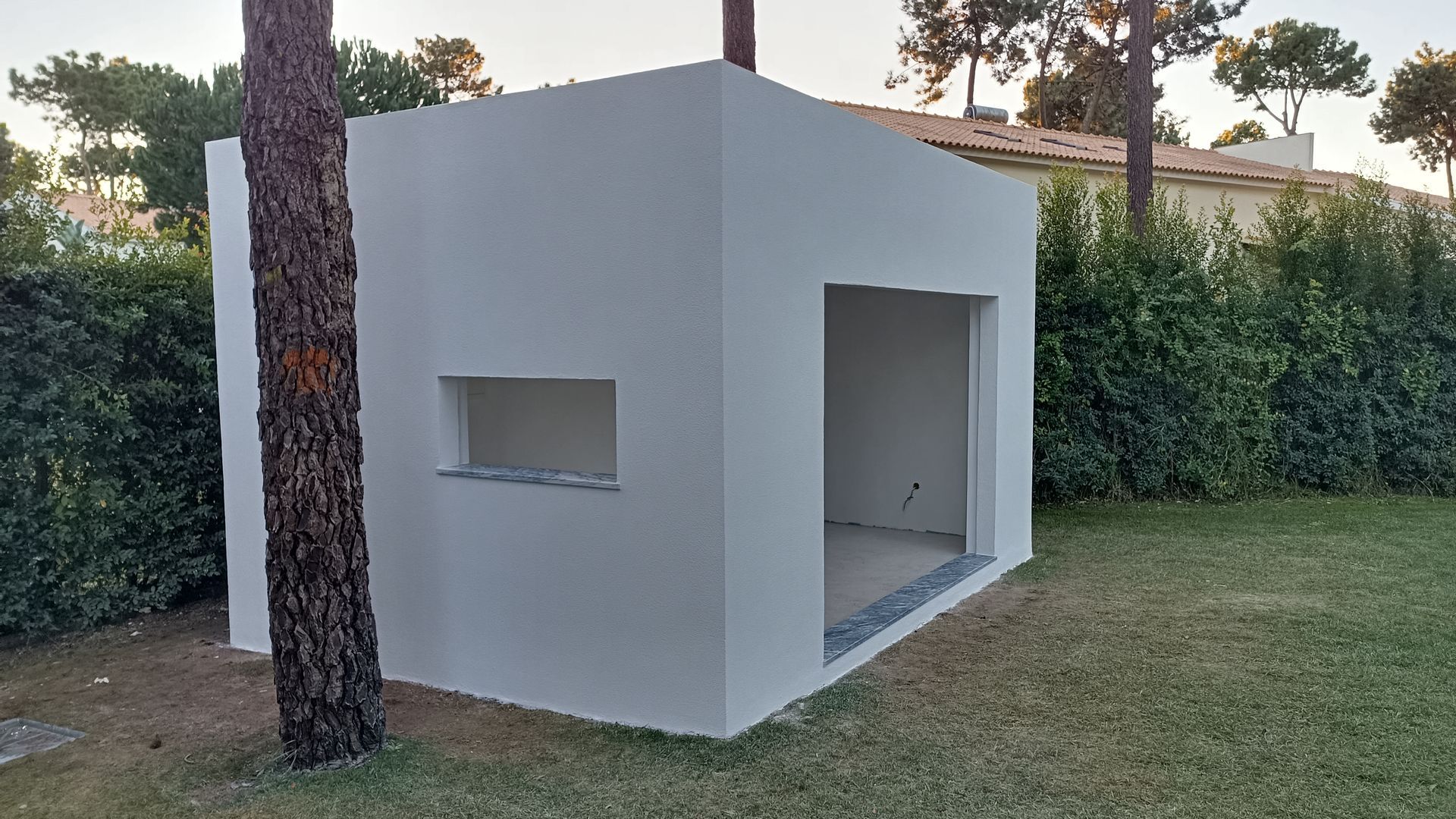 Uma pequena estrutura retangular branca com uma porta aberta e uma janela em um gramado, ao lado de uma árvore.