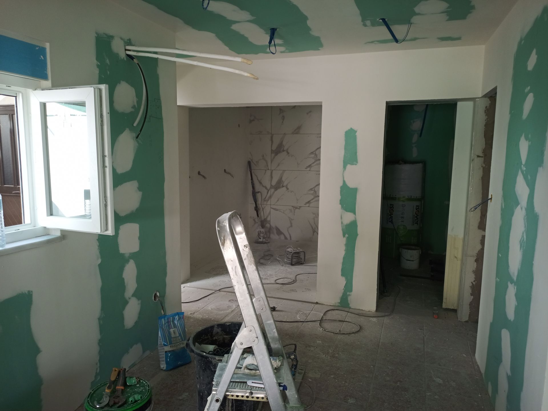Interior de um cômodo em construção, drywall verde, paredes inacabadas, escada, balde, portas abertas, fiação exposta.