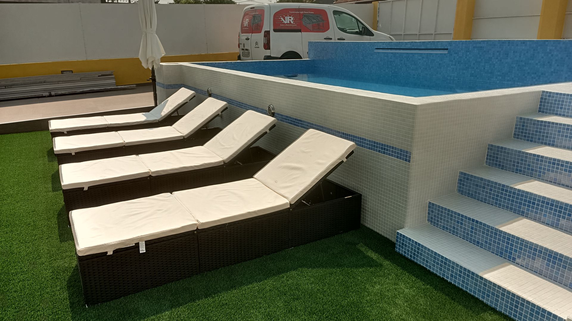 À beira da piscina com espreguiçadeiras em grama sintética. Piscina com azulejos azuis, espreguiçadeiras brancas e uma van ao fundo.
