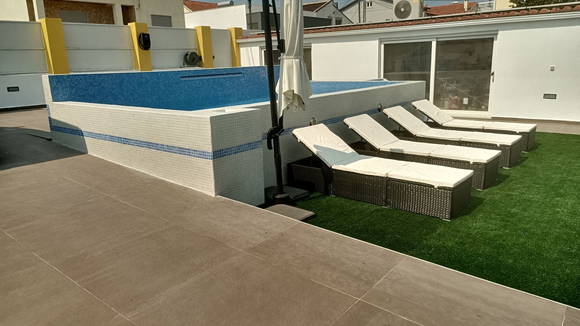 Cena à beira da piscina: piscina retangular com cobertura azul, espreguiçadeiras e grama sintética em um dia ensolarado.