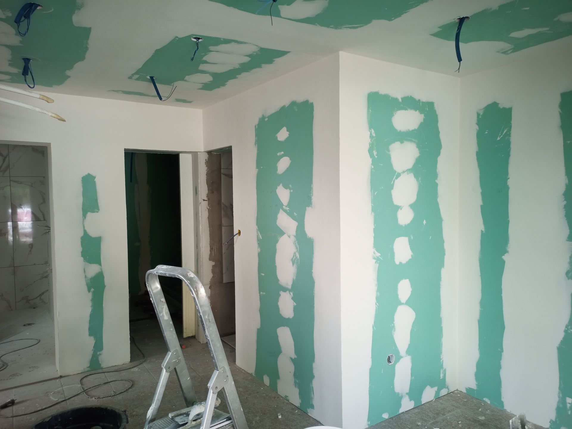 Vista interna de um cômodo em construção com drywall verde e paredes brancas. Uma escada e fiação elétrica são visíveis.