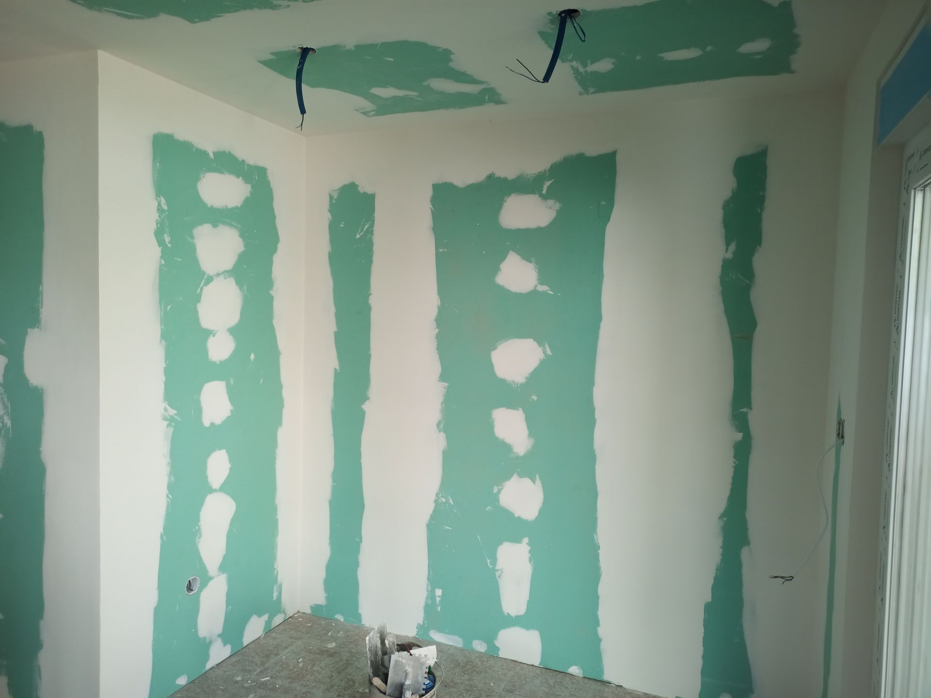 Sala em construção, com drywall verde, paredes brancas e fiação elétrica exposta.