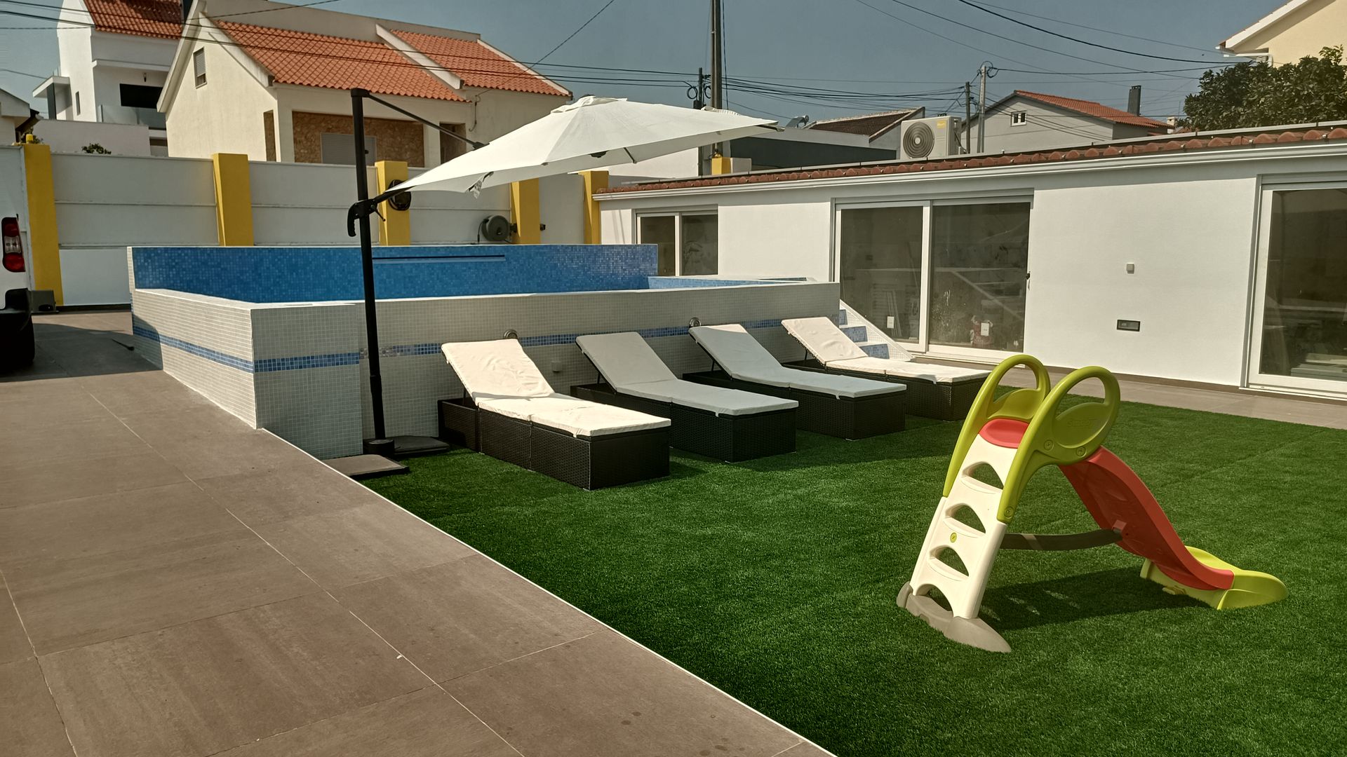 Área da piscina no quintal com espreguiçadeiras, guarda-sol, escorregador e grama sintética.