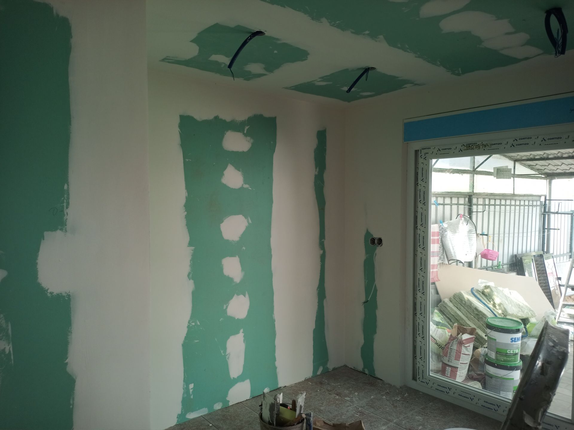 Quarto interior em construção, com drywall verde, paredes brancas e uma janela.