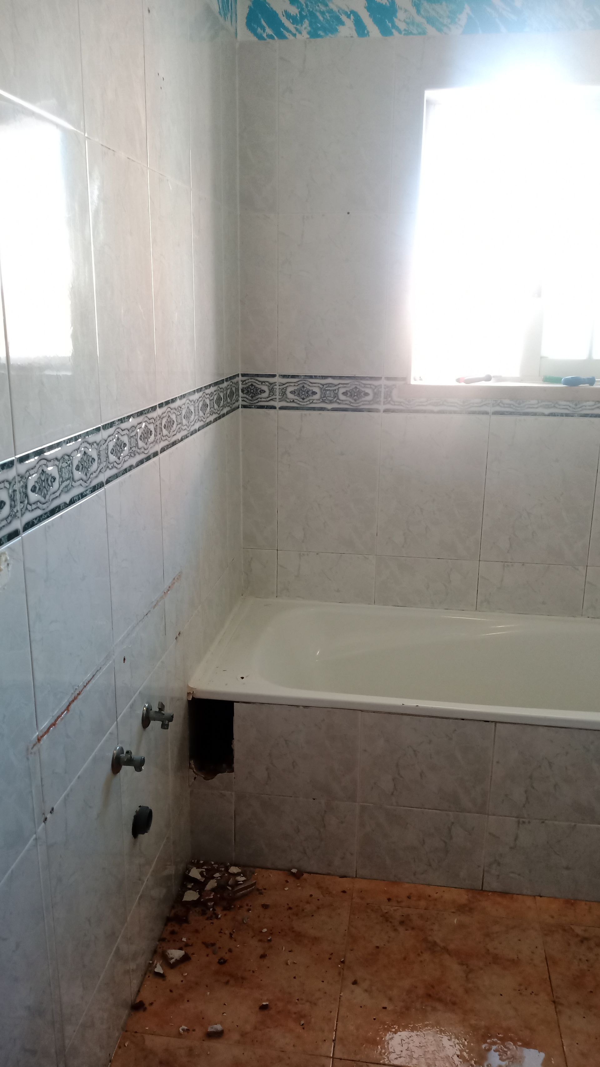 Banheiro com paredes revestidas de azulejos brancos, banheira e janela. O piso marrom é visível.