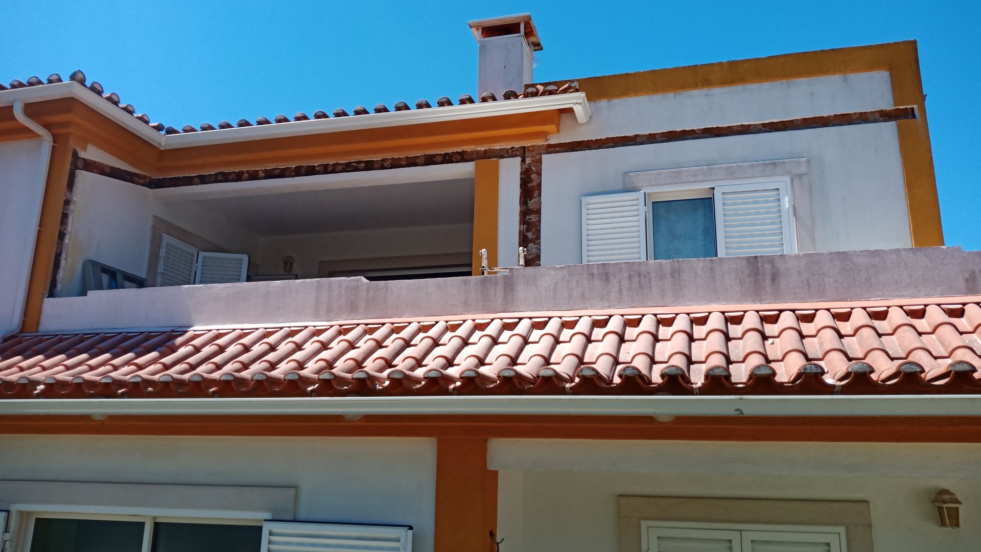 Casa branca com detalhes e telhado laranja, varanda e céu azul.