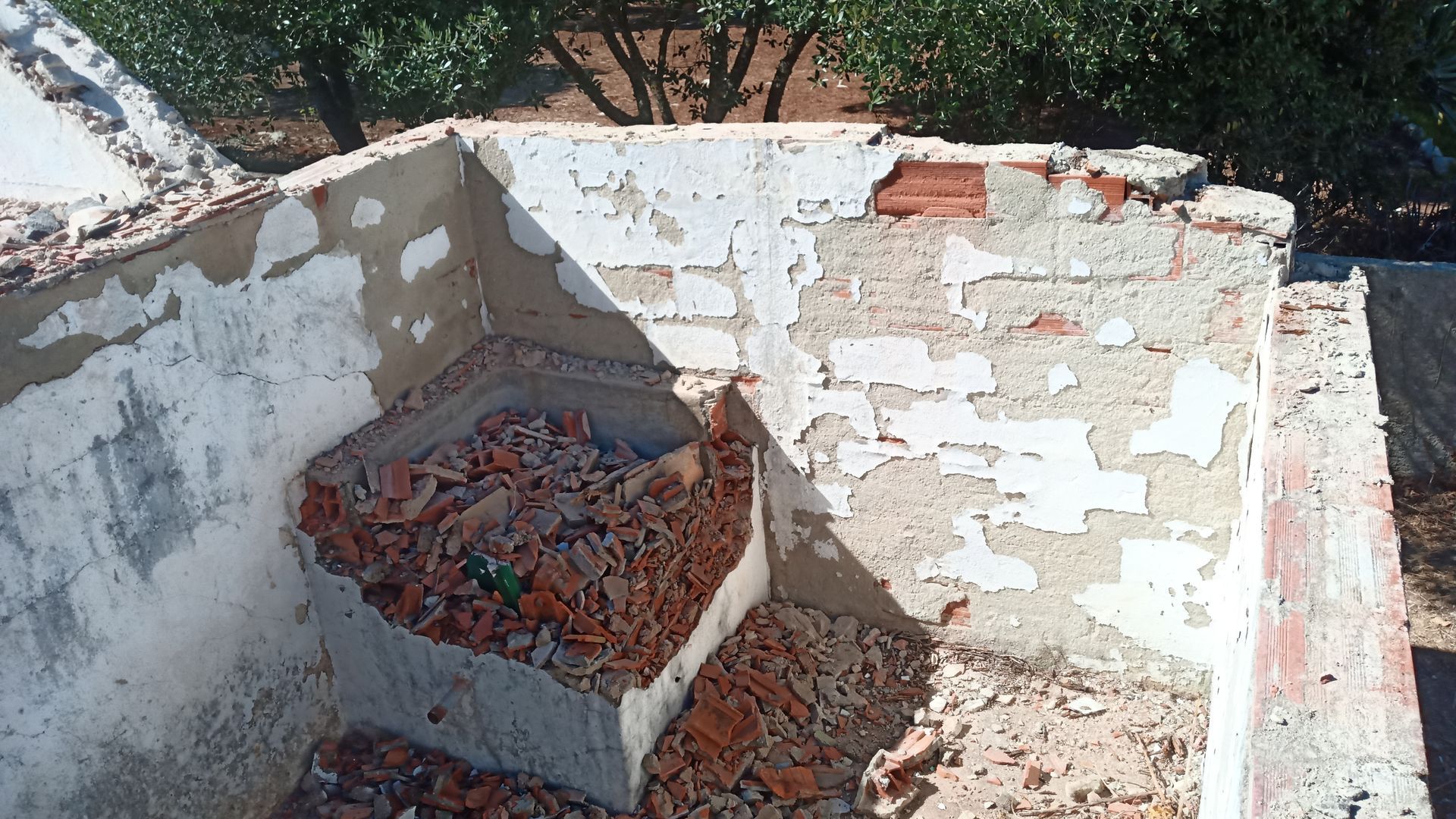 Estrutura danificada de tijolo e concreto com tinta descascando. Entulho no interior. Ambiente externo ensolarado.