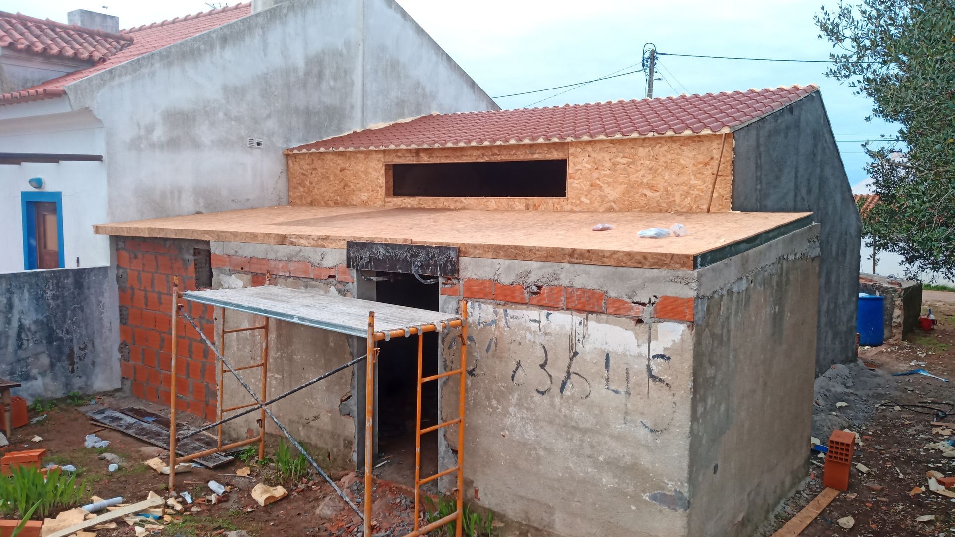 Construção de um pequeno edifício de concreto. Andaimes, estrutura de madeira e telhado de telhas vermelhas visíveis.