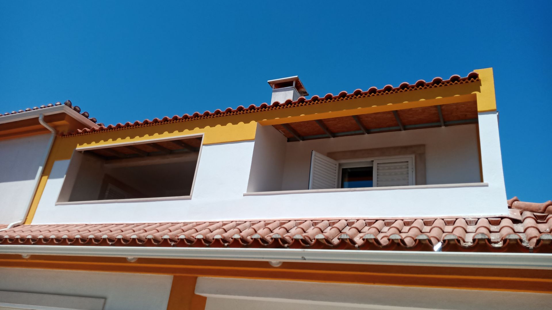 Prédio branco e amarelo com telhas laranja, duas janelas e céu azul.