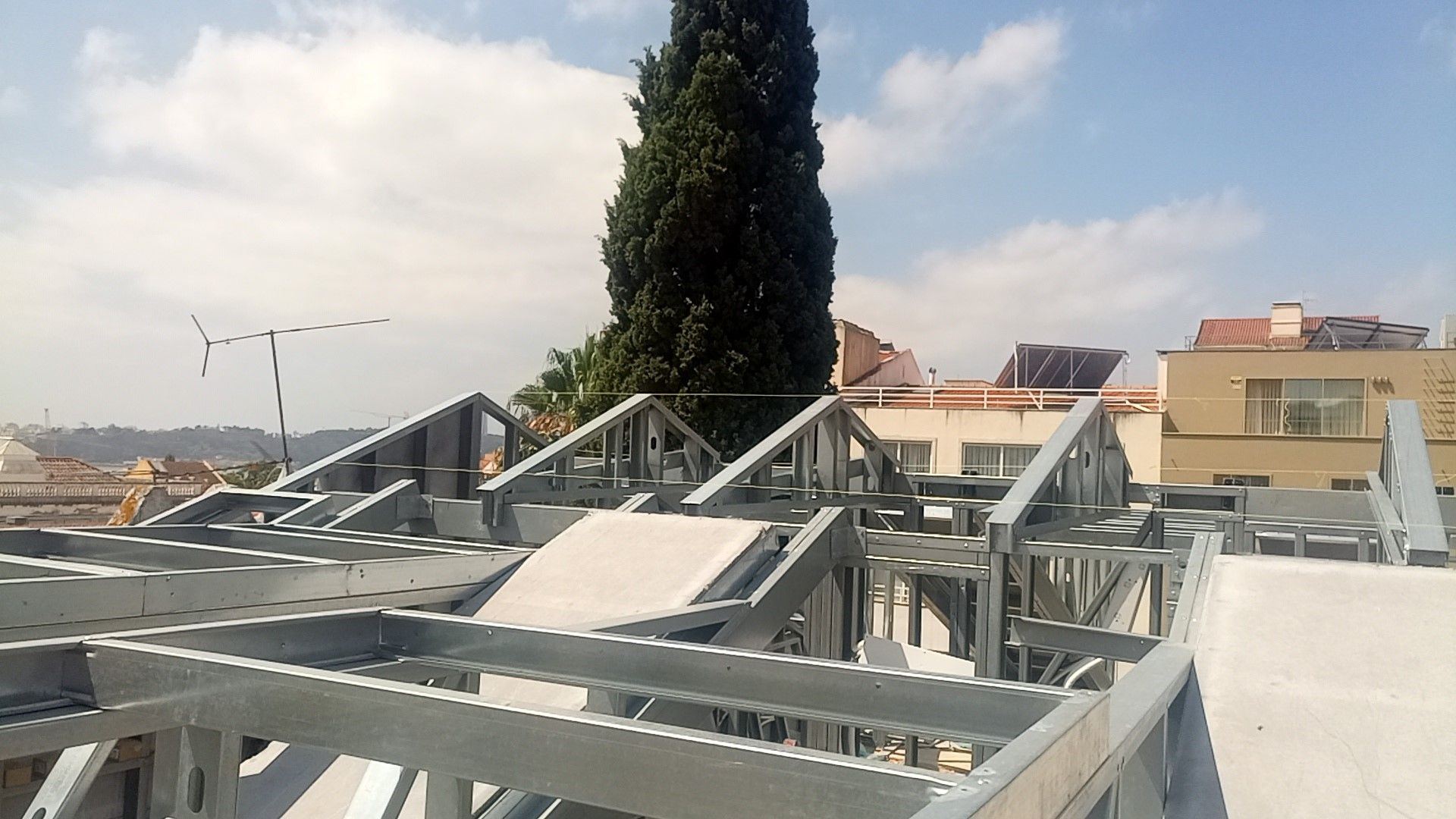 Estrutura metálica de um telhado em construção contra um céu azul com uma árvore alta ao fundo.