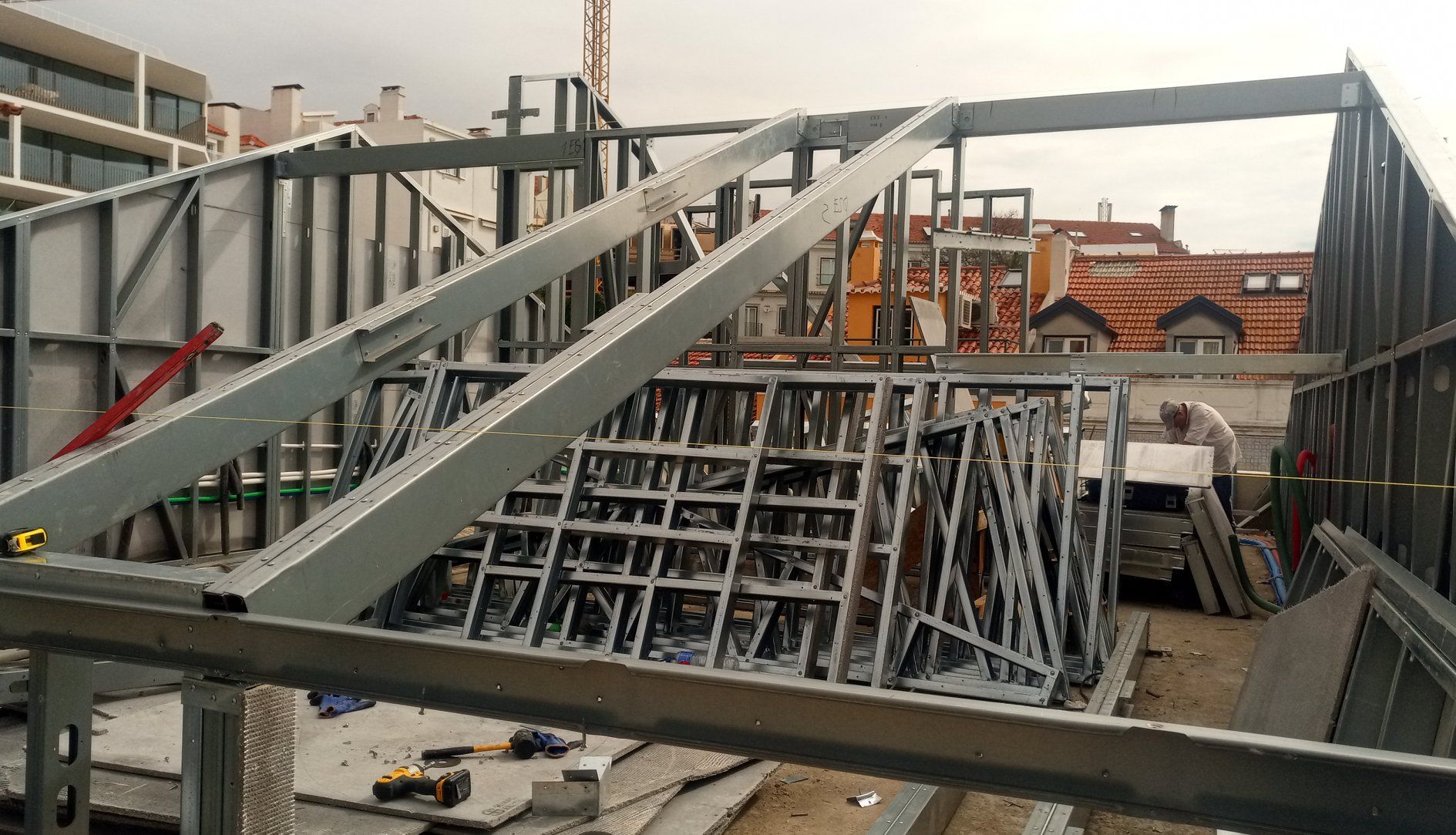 Canteiro de obras com estrutura metálica. Prédios e guindaste ao fundo.